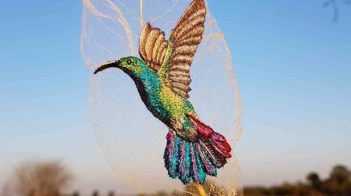 A highly detailed, iridescent Hummingbird (Beija-flor) embroidered on a translucent skeletonized leaf, held up against a blue sky. This image illustrates the Semiotic Paradox of capturing a creature of hyper-metabolism on the medium of stasis.