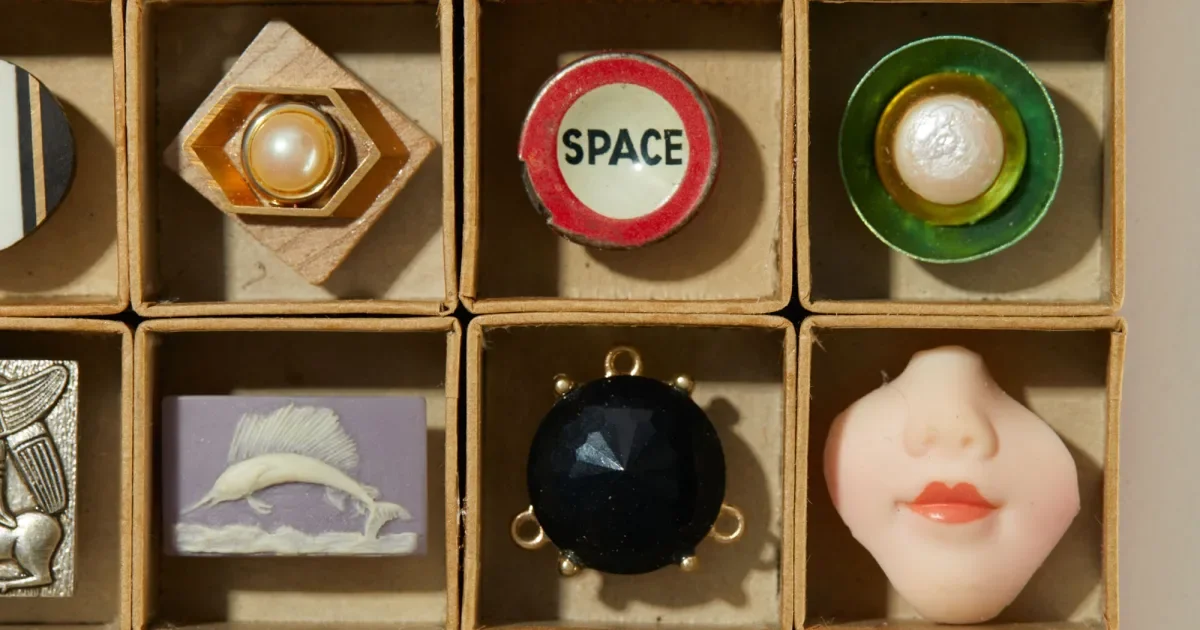 A detailed view of small found objects arranged in a box, including buttons, a porcelain doll face, and vintage trinkets, illustrating Robert Ebendorf’s use of reclaimed materials in jewelry design.