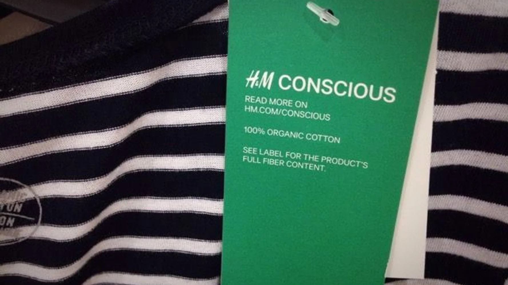 A close-up image of a vibrant green H&M "Conscious" hang tag on a striped shirt. The tag reads "100% Organic Cotton" and directs the user to "Read more on HM.com/conscious," illustrating a visible, consumer-facing attempt at sustainability marketing.