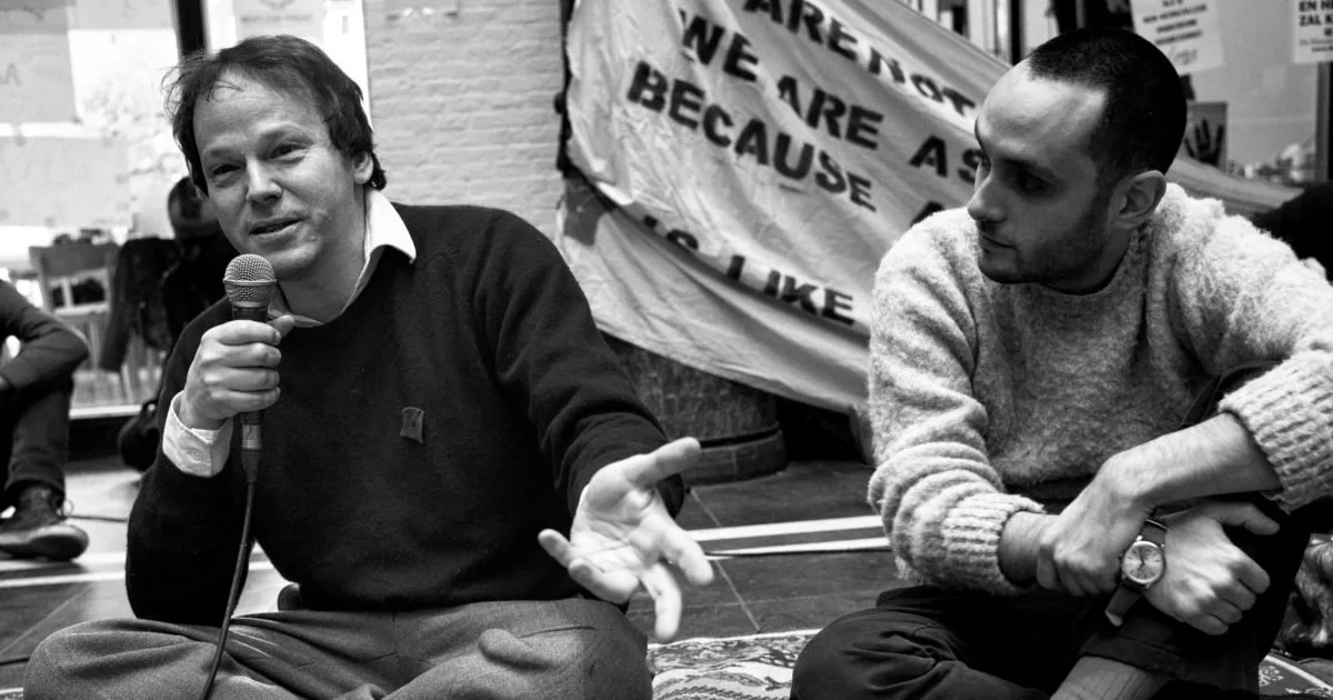 A black and white photo of anthropologist David Graeber speaking into a microphone while sitting on the floor, engaging in a discussion about social and economic theory.