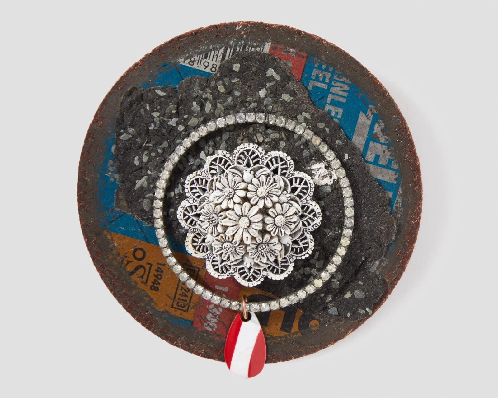 Close-up of Robert Ebendorf’s "Cutting Disc Brooch," an assemblage of a rusted industrial metal cutting disc, a central white plastic floral motif, rhinestone chains, and a red and white plastic tag, showcasing his found object jewelry philosophy.