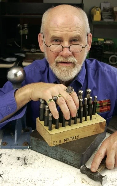 Portrait of Robert Ebendorf, wearing glasses and a blue shirt, leaning on a workbench with his hand resting on a set of metalsmithing ring mandrels labeled "ECU METALS."