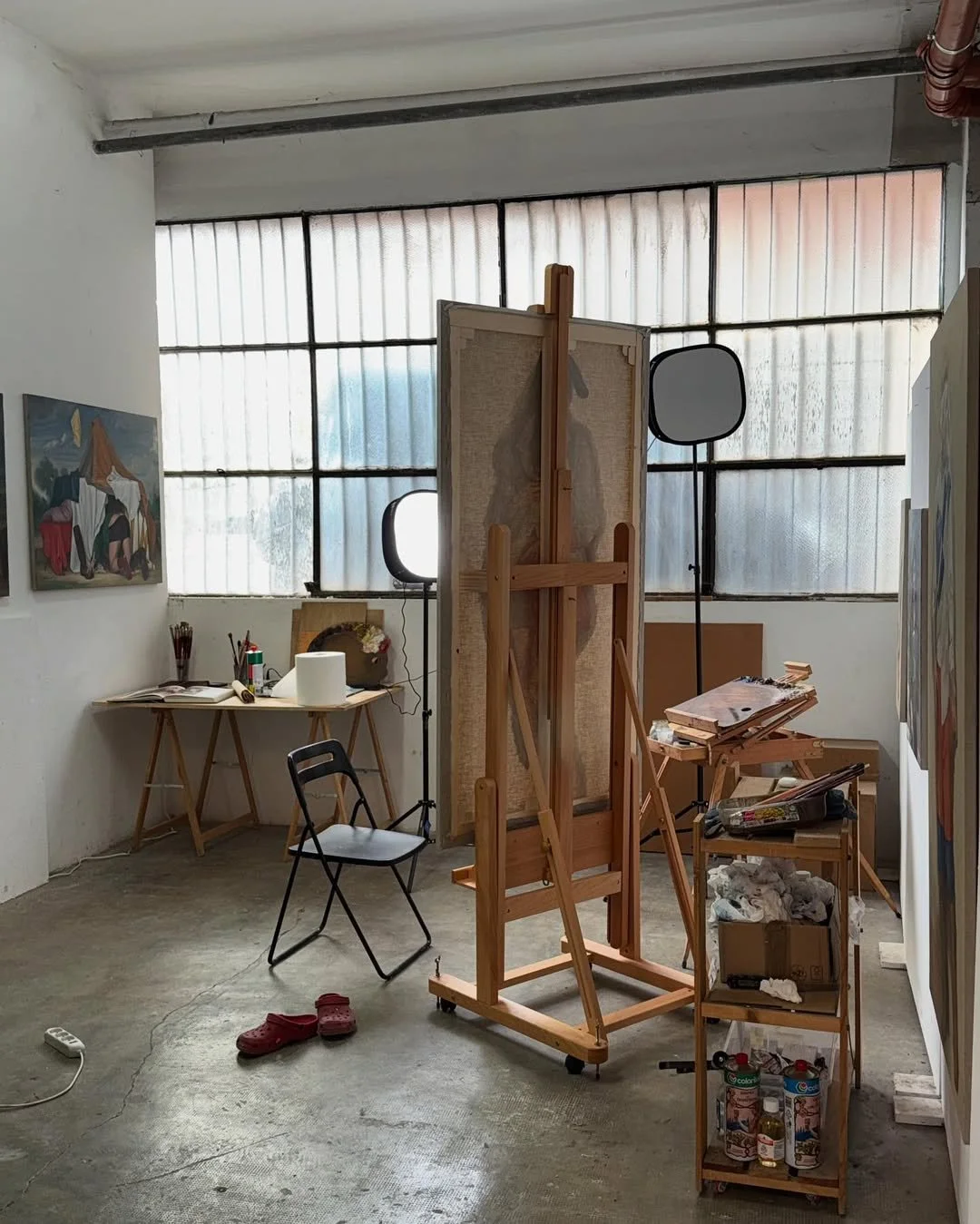 An artist's studio with high industrial windows, featuring a wooden easel with a canvas in progress, studio lights, and a small table with art supplies, representing the workspace of Nazar Strelyaev-Nazarko.