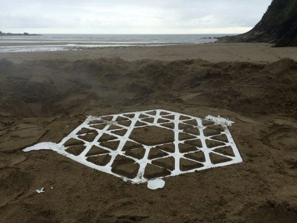 The molten pewter geometric structure of Max Lamb's Pewter Stool cooling directly in a hand-carved sand mold on Caerhays Beach under an overcast sky.