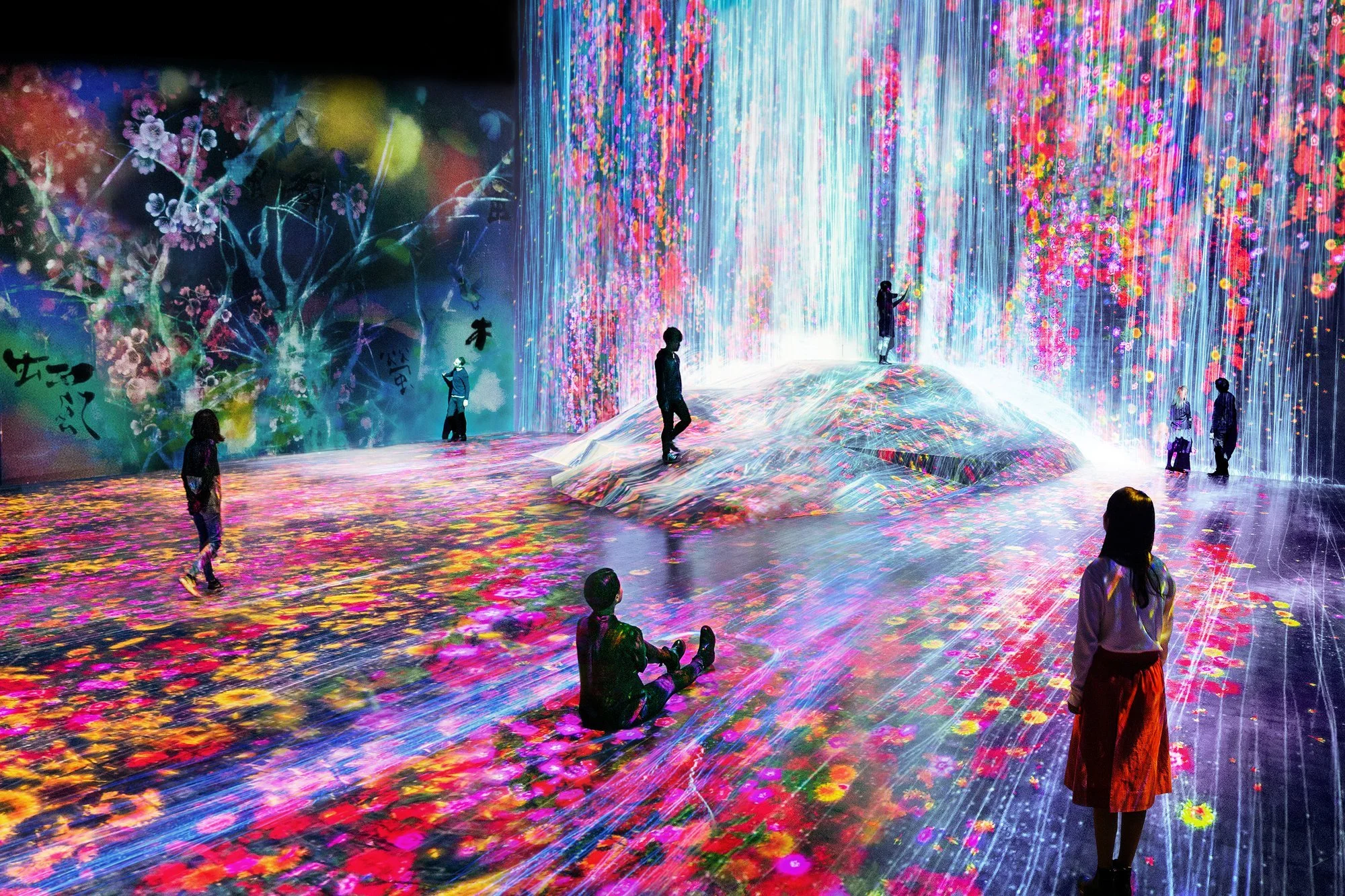 An immersive digital art installation by teamLab inside Superblue, featuring a large illuminated artificial hill with a projected cascading waterfall of light amidst a field of vibrant digital flowers, with human figures interacting in the space.