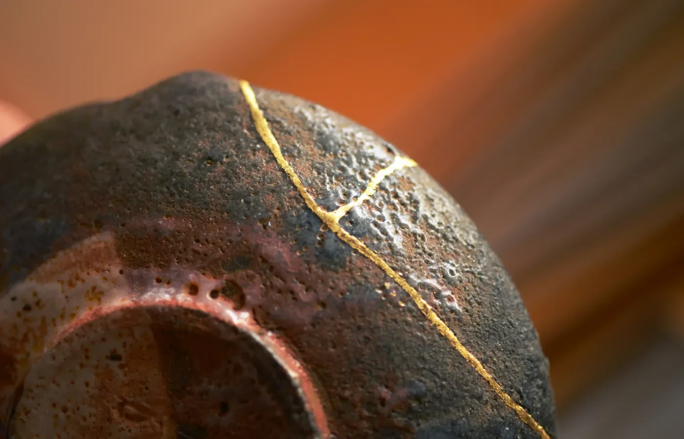 A close-up macro shot of a dark, textured ceramic bowl repaired with kintsugi, showing the golden lacquer celebrating the crack as part of its history.