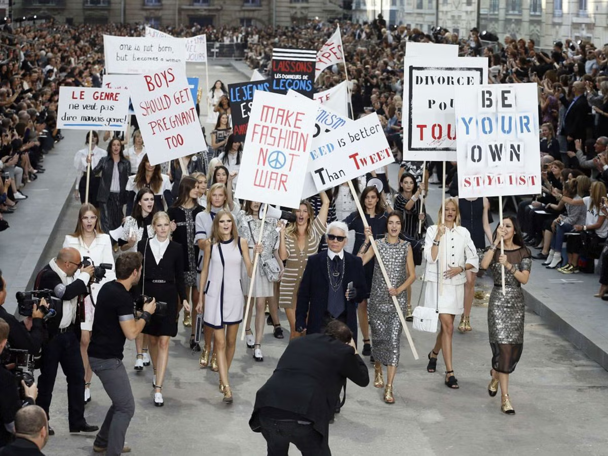 Models led by Karl Lagerfeld in the Chanel Spring/Summer 2015 runway show, holding faux protest signs, illustrating the commodification of dissent in high fashion.