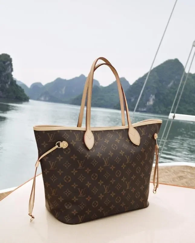 A Louis Vuitton Neverfull monogram tote bag on a boat deck with a scenic coastal background, illustrating the concept of a Hollowed Object that relies on logo recognition rather than semantic depth.