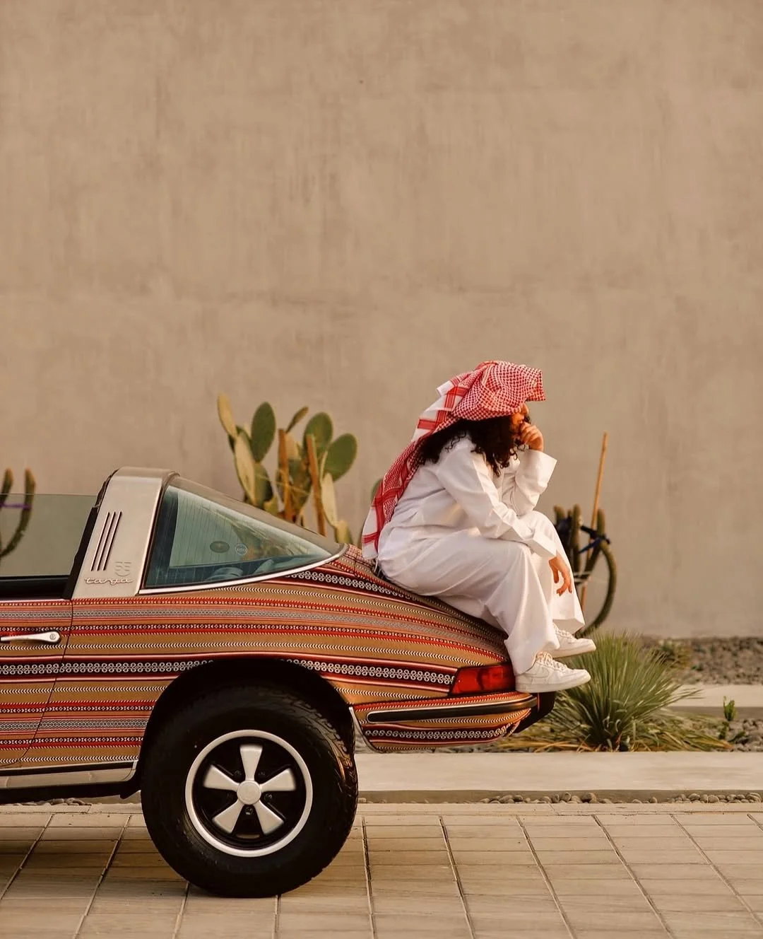 The finished custom-painted classic Porsche 911 Targa adorned entirely in fine Al Sadu geometric livery stripes, parked outdoors against a minimalist concrete wall with a person in traditional Gulf attire sitting on the rear decklid.