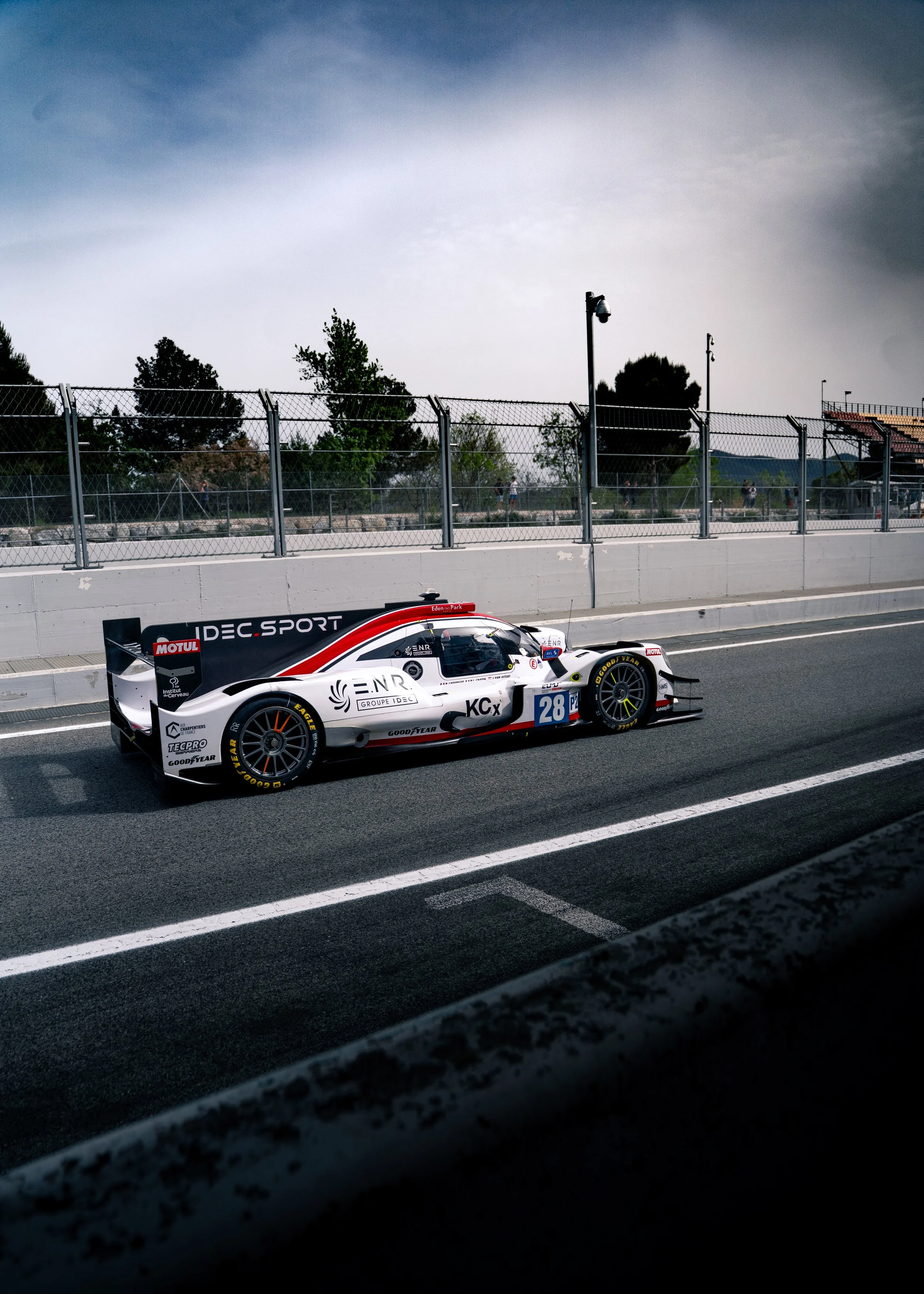A race car on a race track with a white barrier wall, chain-link fence, trees, and cloudy sky in the background.