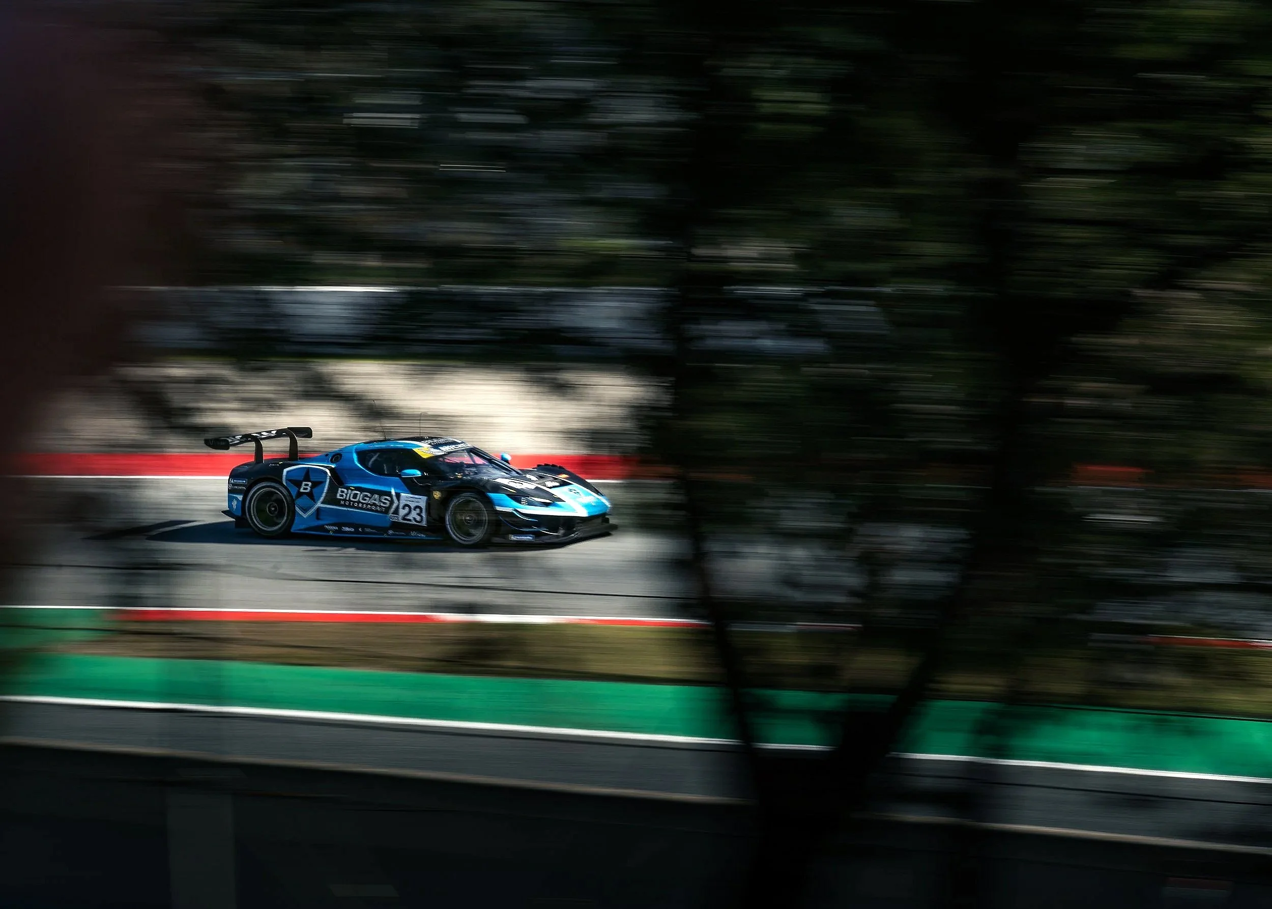 A blue race car with a black spoiler and sponsorship logos, including 'BioGas Motorsport' and '23,' speeding along a race track with a blurred background of trees and trackside barriers.