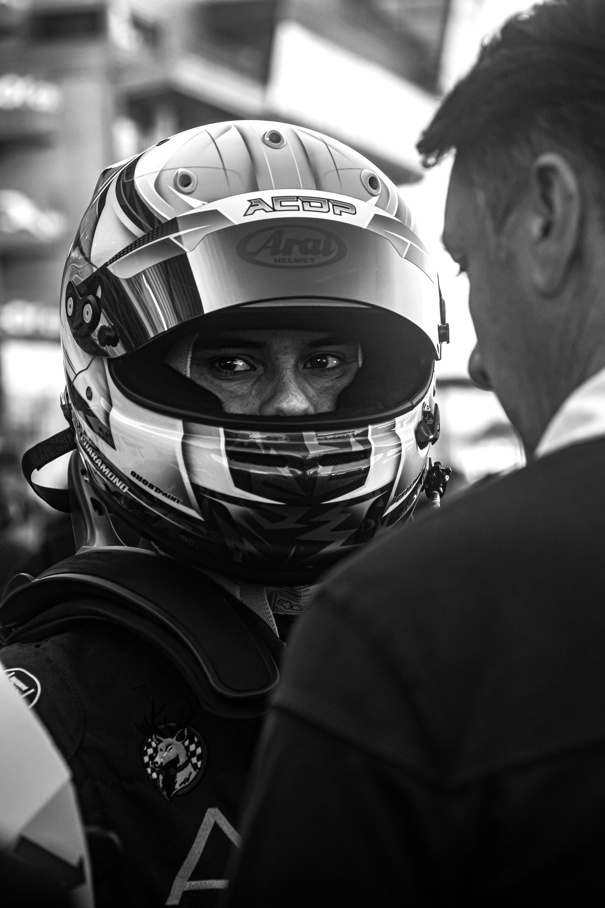A race car driver wearing a helmet and racing suit, talking to a man in a busy or pit area.