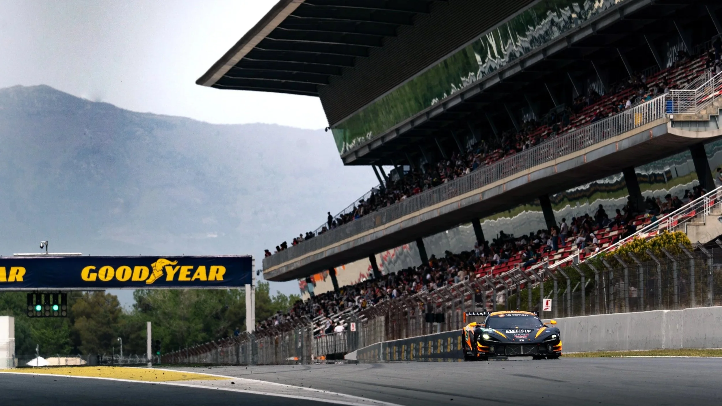 Race car on track with grandstands filled with spectators at a racing event, with mountains and cloudy sky in background.