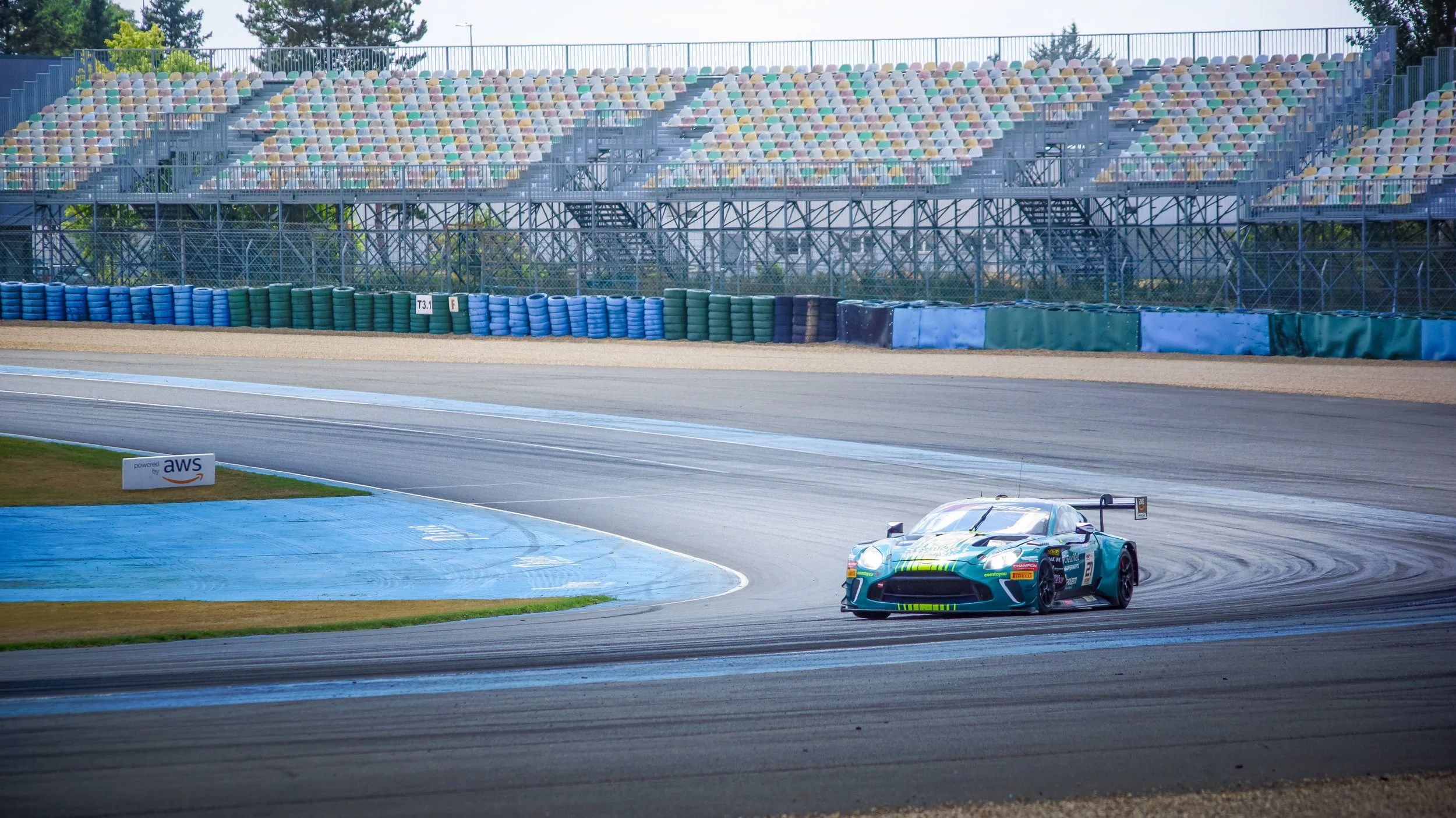 A race car on a racetrack with empty grandstands in the background and a AWS sign on a small billboard near the track.