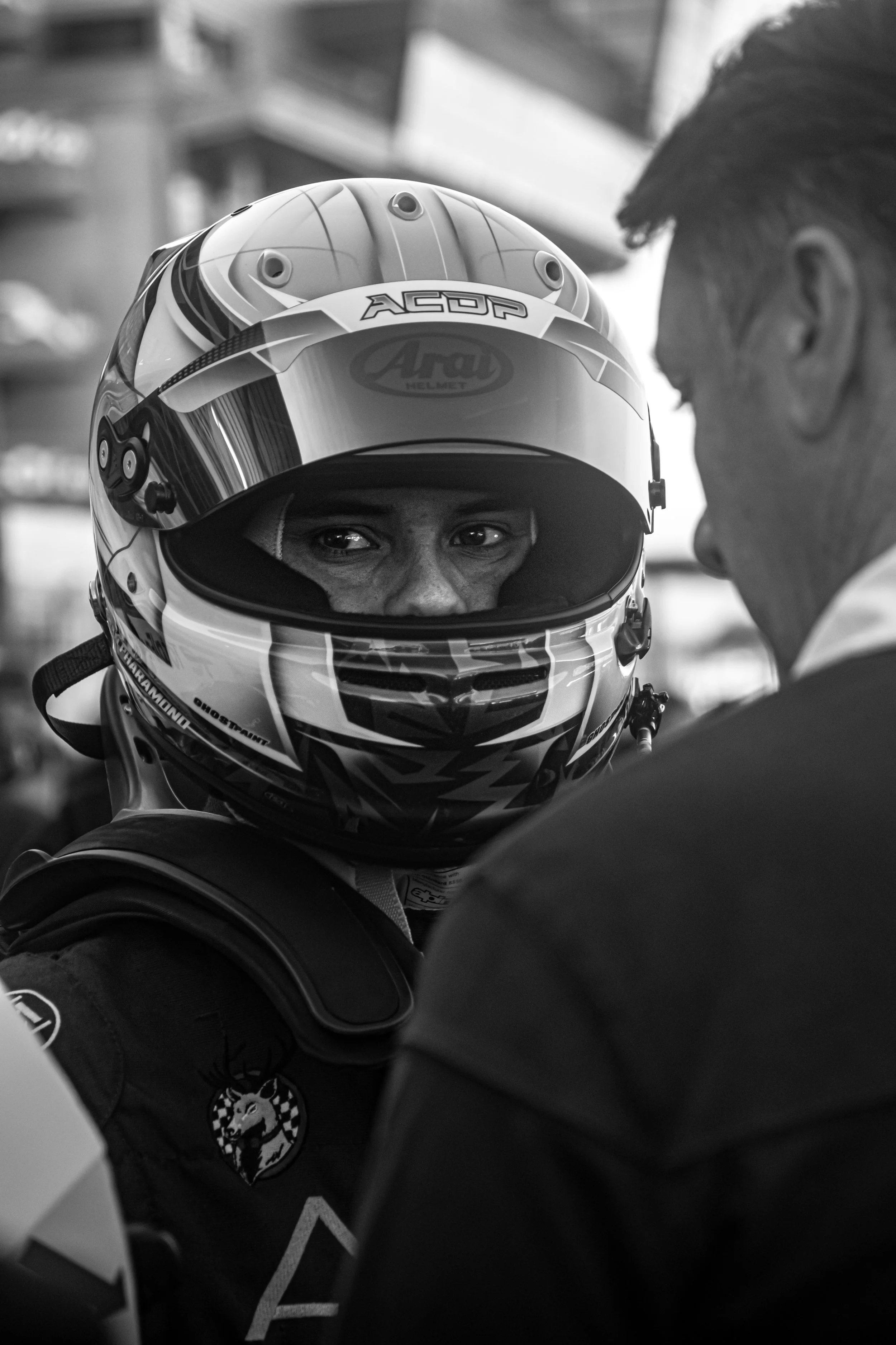 A race car driver wearing a racing helmet and suit, looking at a person, in black and white.