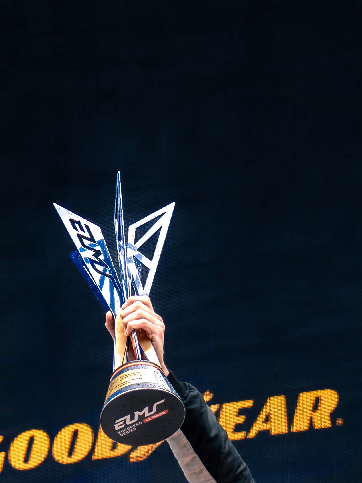 A person holding a trophy with the logo of the European Le Mans Series and the number 6, celebrating on a stage with a black background and yellow illuminated text.