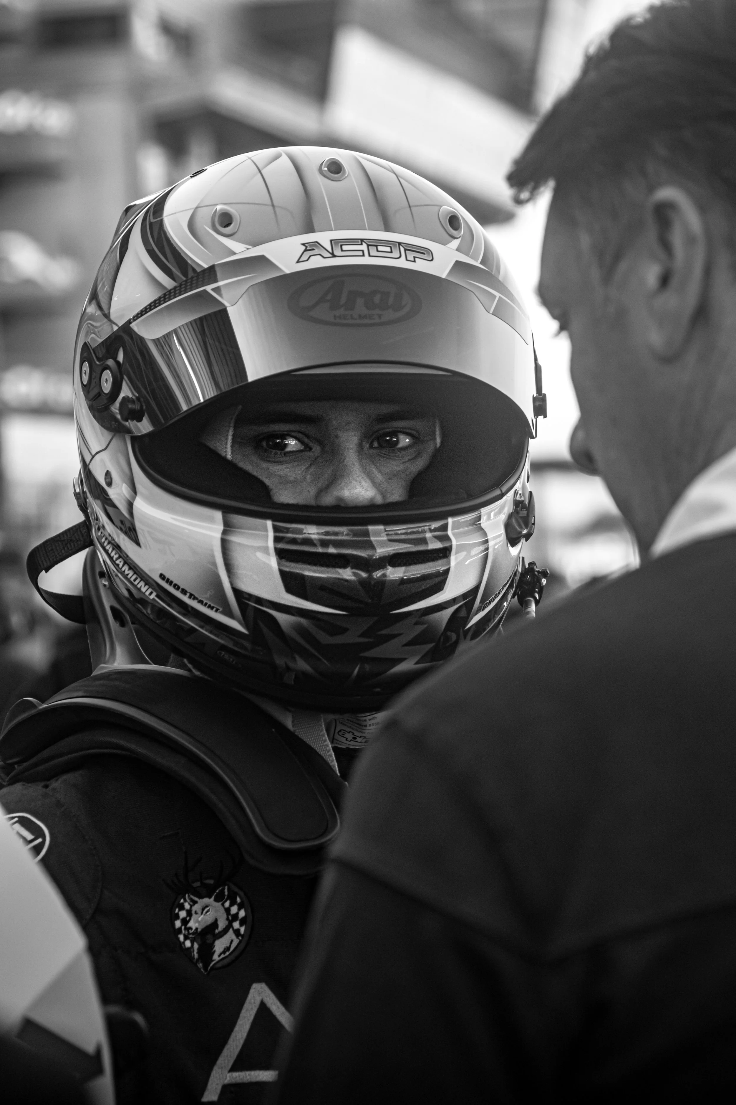 A race car driver wearing a helmet and racing suit, talking to a man in a busy or pit area.