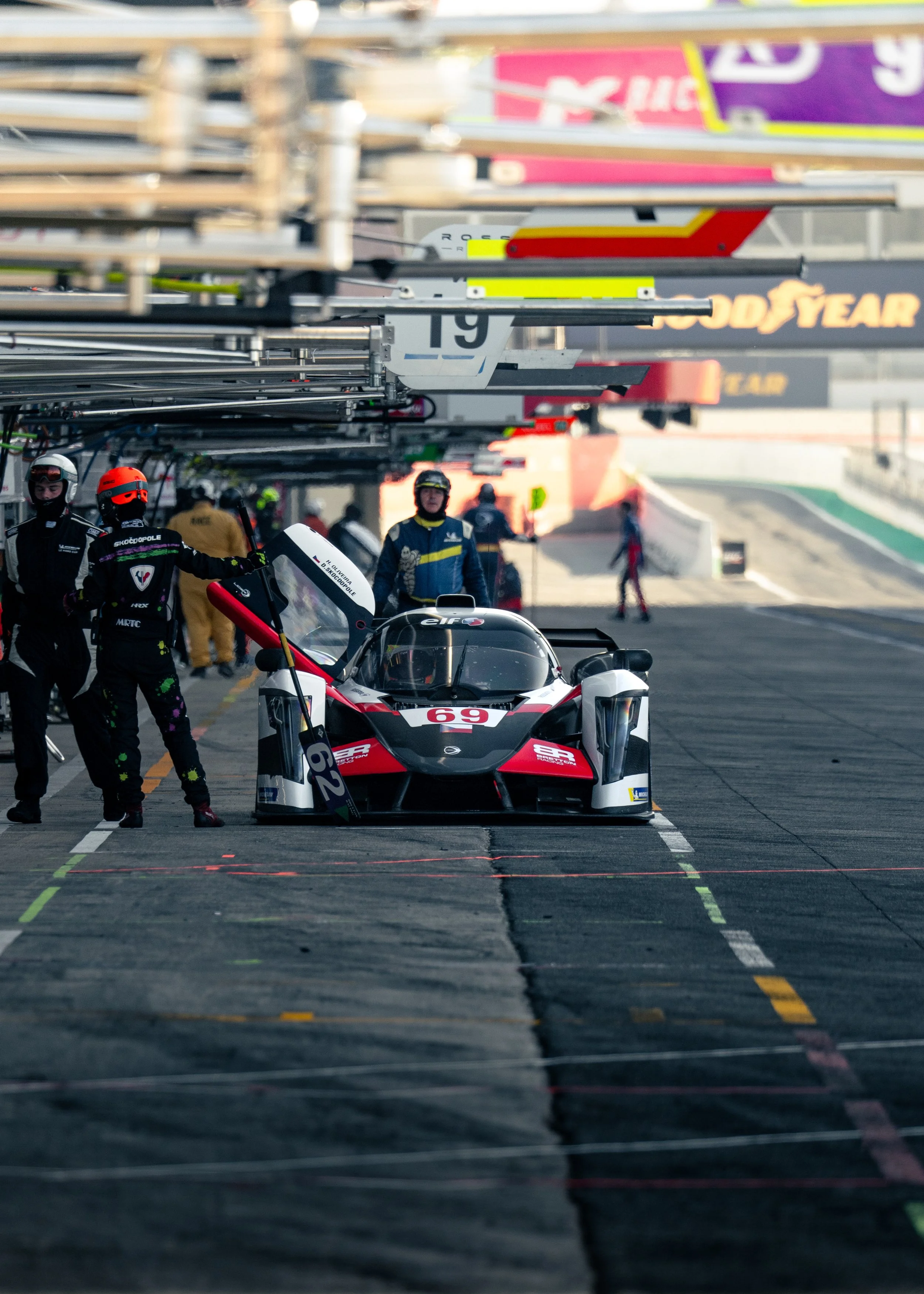 A race car with the number 69 in the pit lane of a race track, with crew members and other personnel around, and equipment overhead.