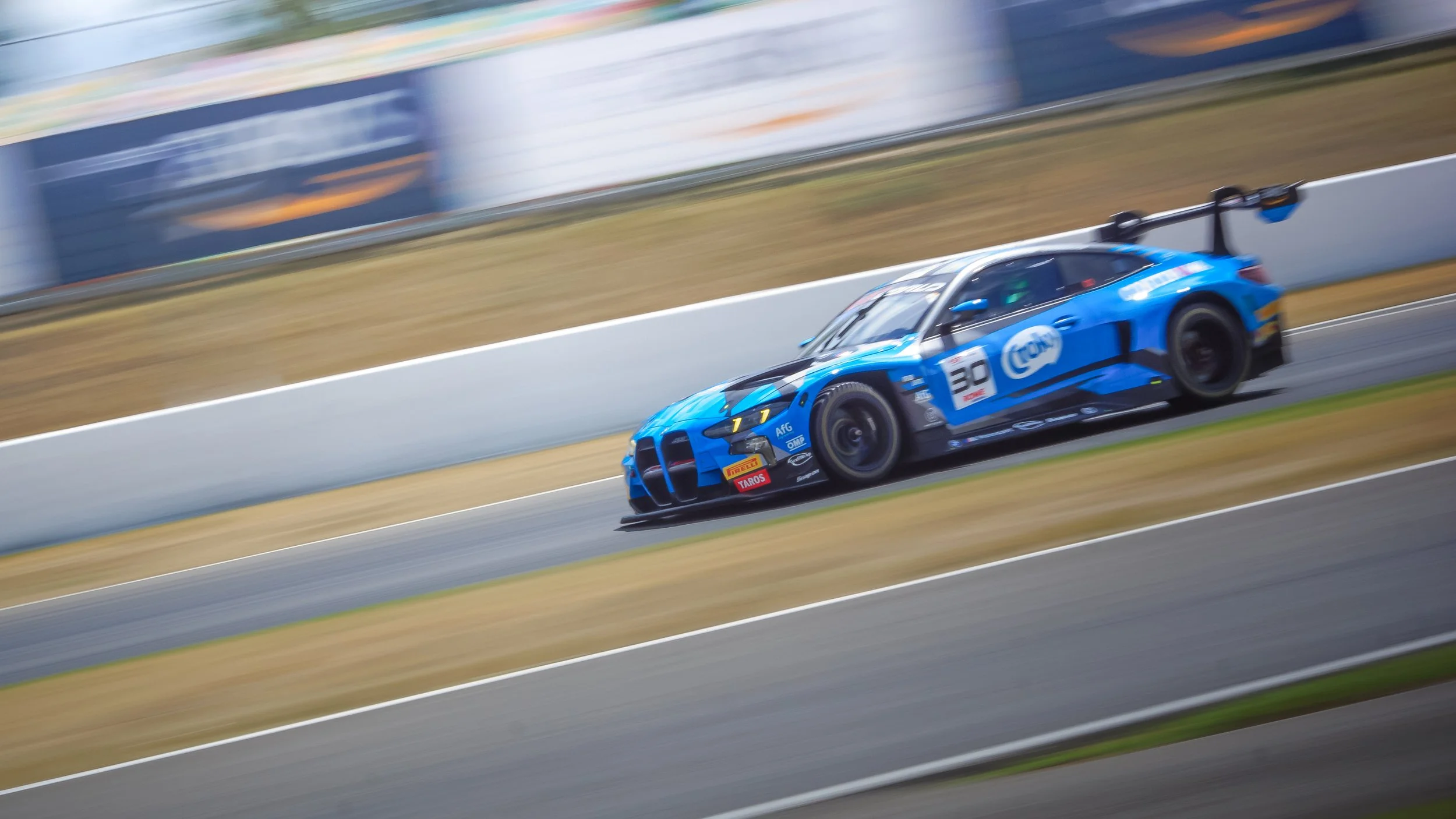 A blue race car with the number 30 on the side speeds along a race track, with motion blur indicating high speed, and a white barrier wall beside the track.