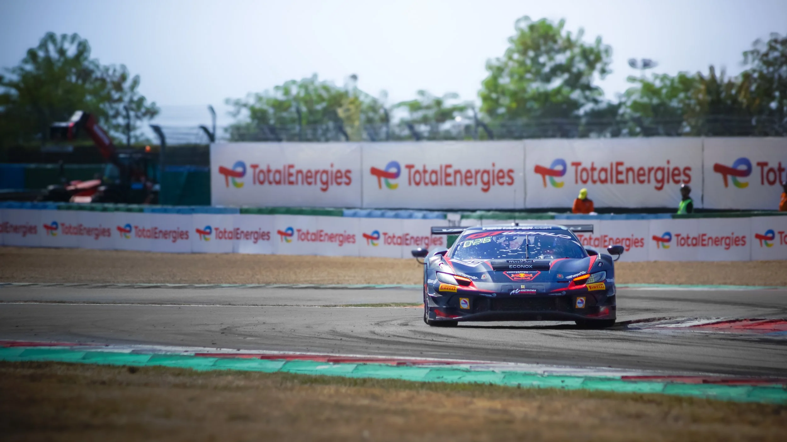 A race car on a racing track approaching a corner, with banners displaying 'TotalEnergies' in the background.