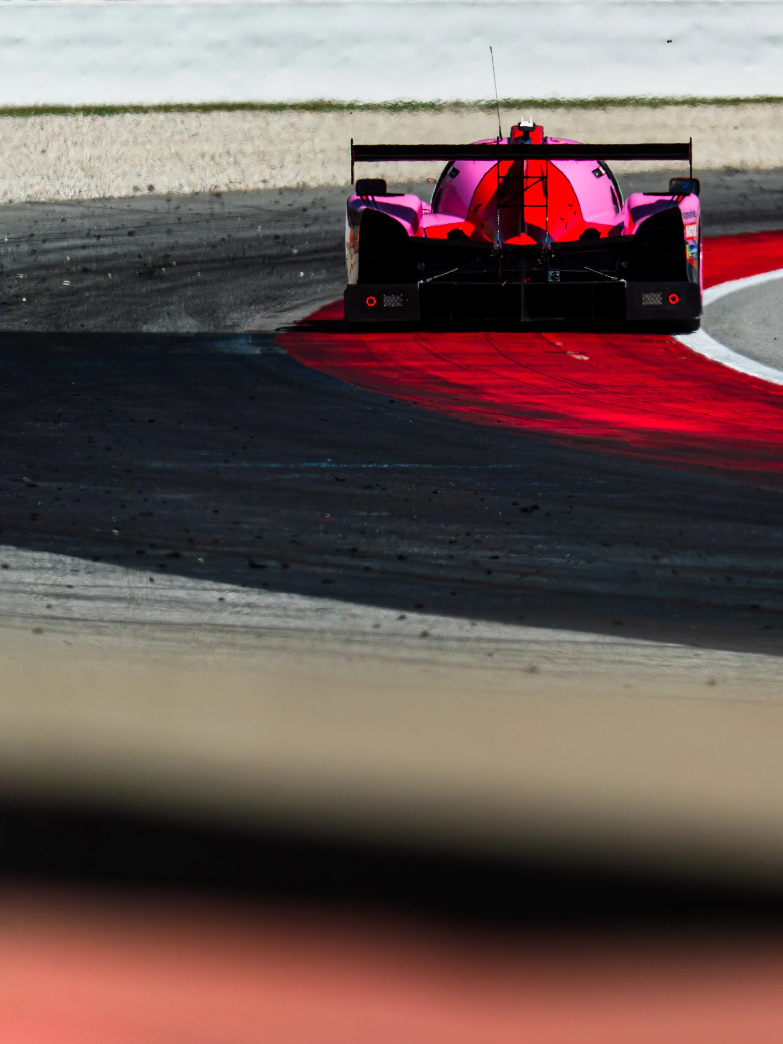 A pink and black race car on a racetrack, viewed from front, with a rear wing and antenna on top.