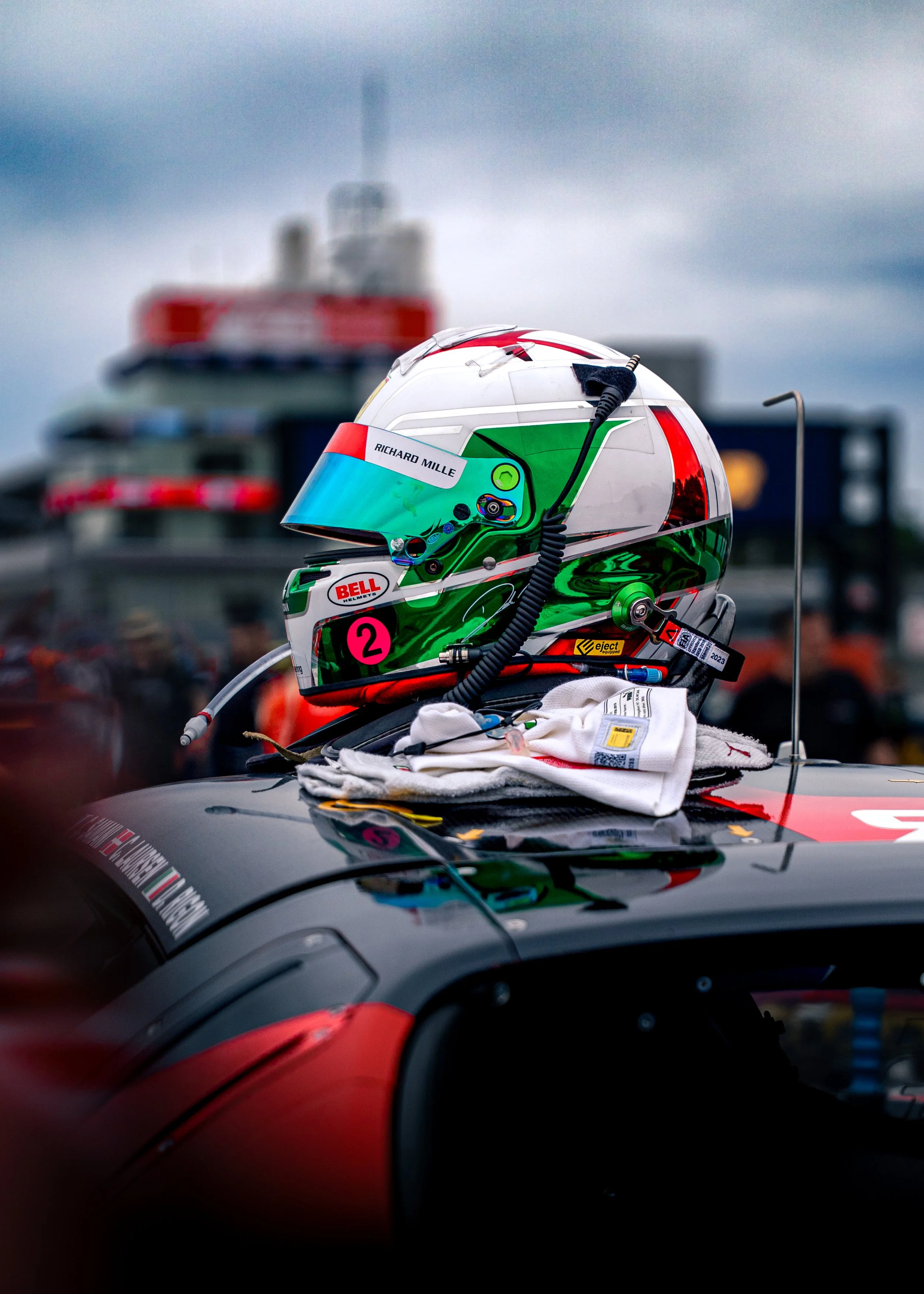 A racing helmet with green, white, and red colors and sponsor logos, placed on the roof of a black race car with a red stripe. The helmet has a visor, audio equipment, and a name tag reading 'Richard Mille.' The background shows a blurred racing scen