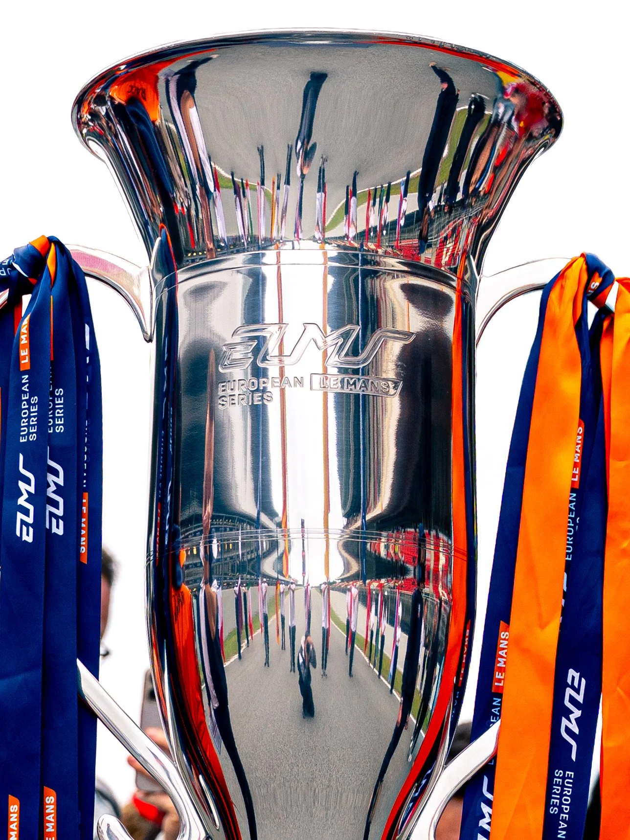 A reflective silver trophy with engraved text and several orange and blue ribbons attached to its handles, displayed at a sporting event.