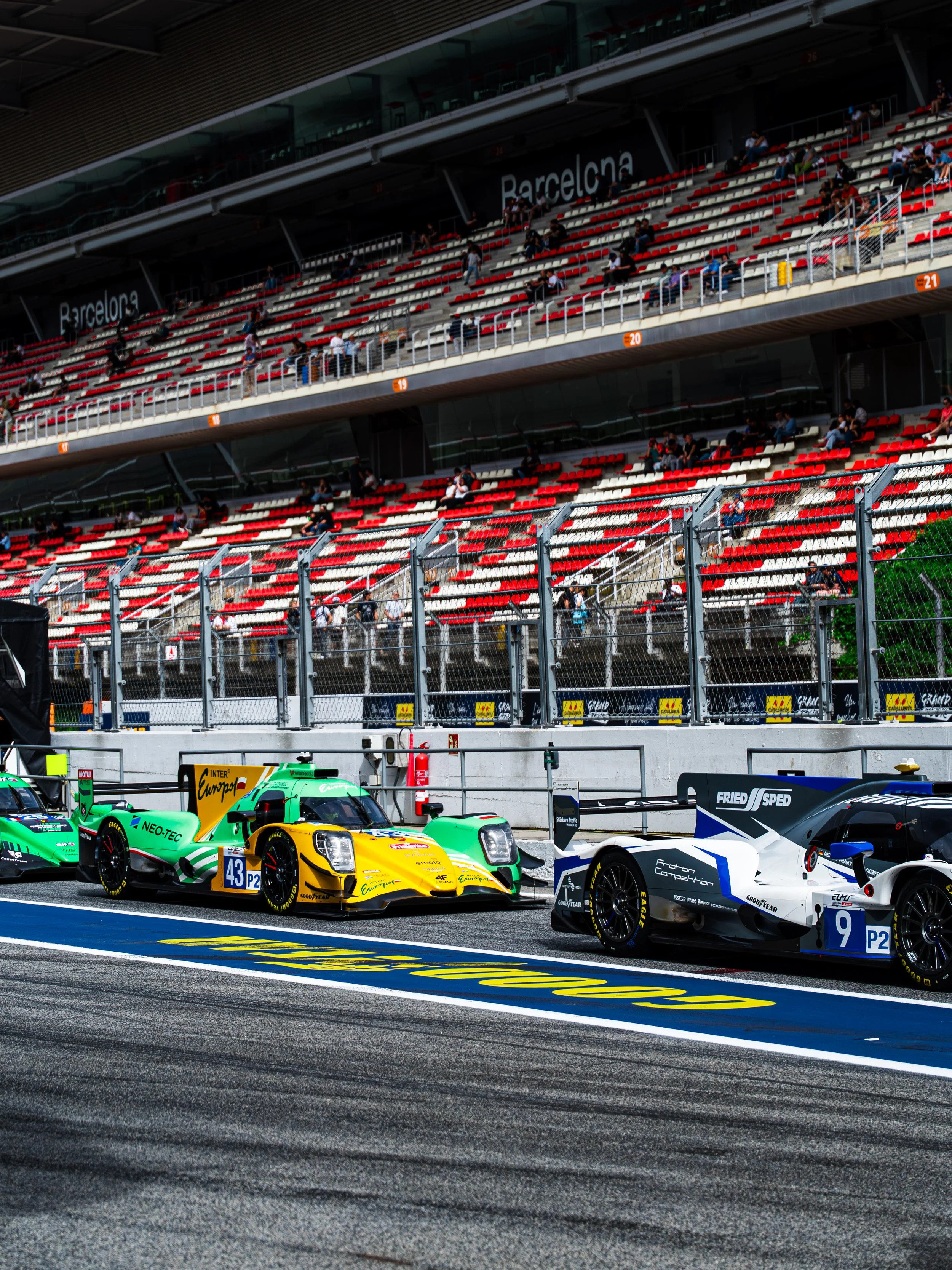 Race cars on a racetrack in front of tiered grandstands at Barcelona circuit, with people seated in the stands and a metal fence in the foreground.