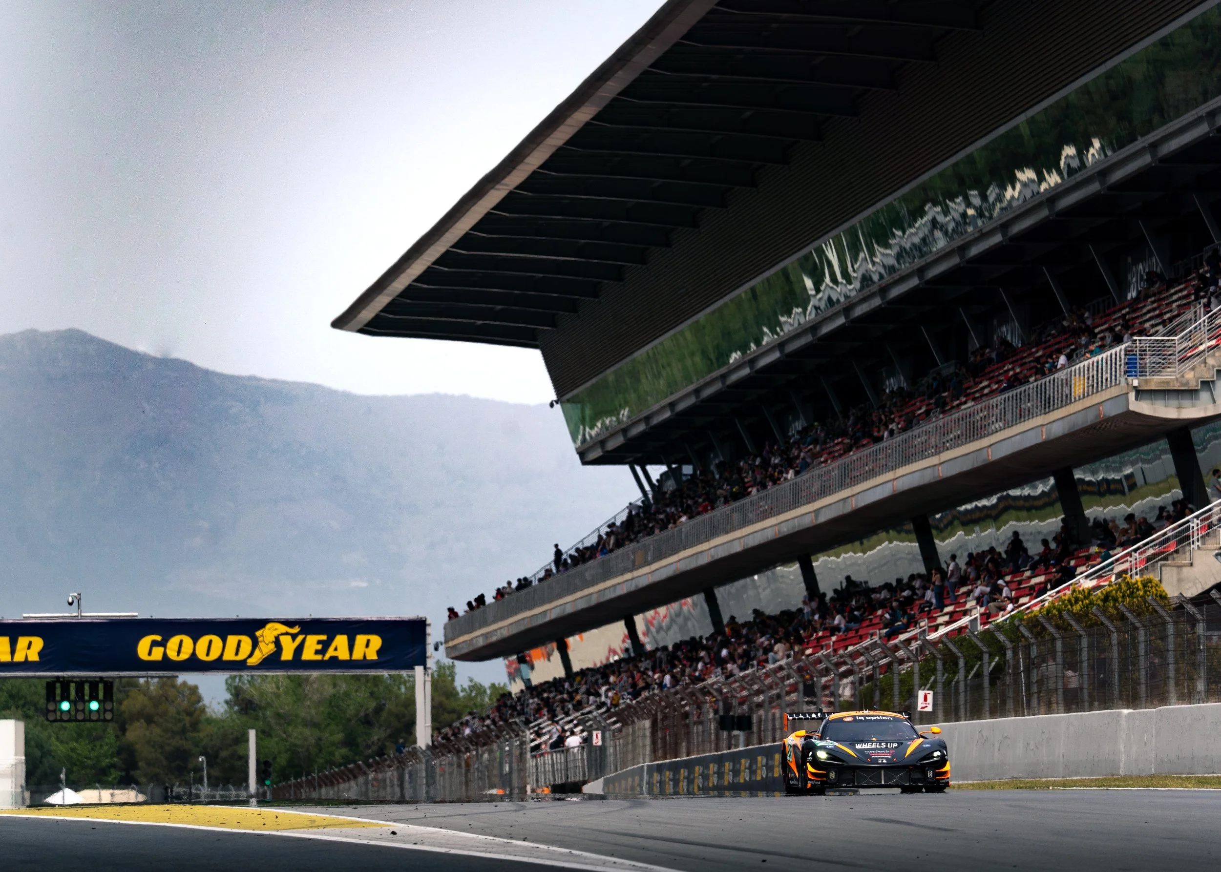 Race car on track with grandstands filled with spectators at a racing event, with mountains and cloudy sky in background.