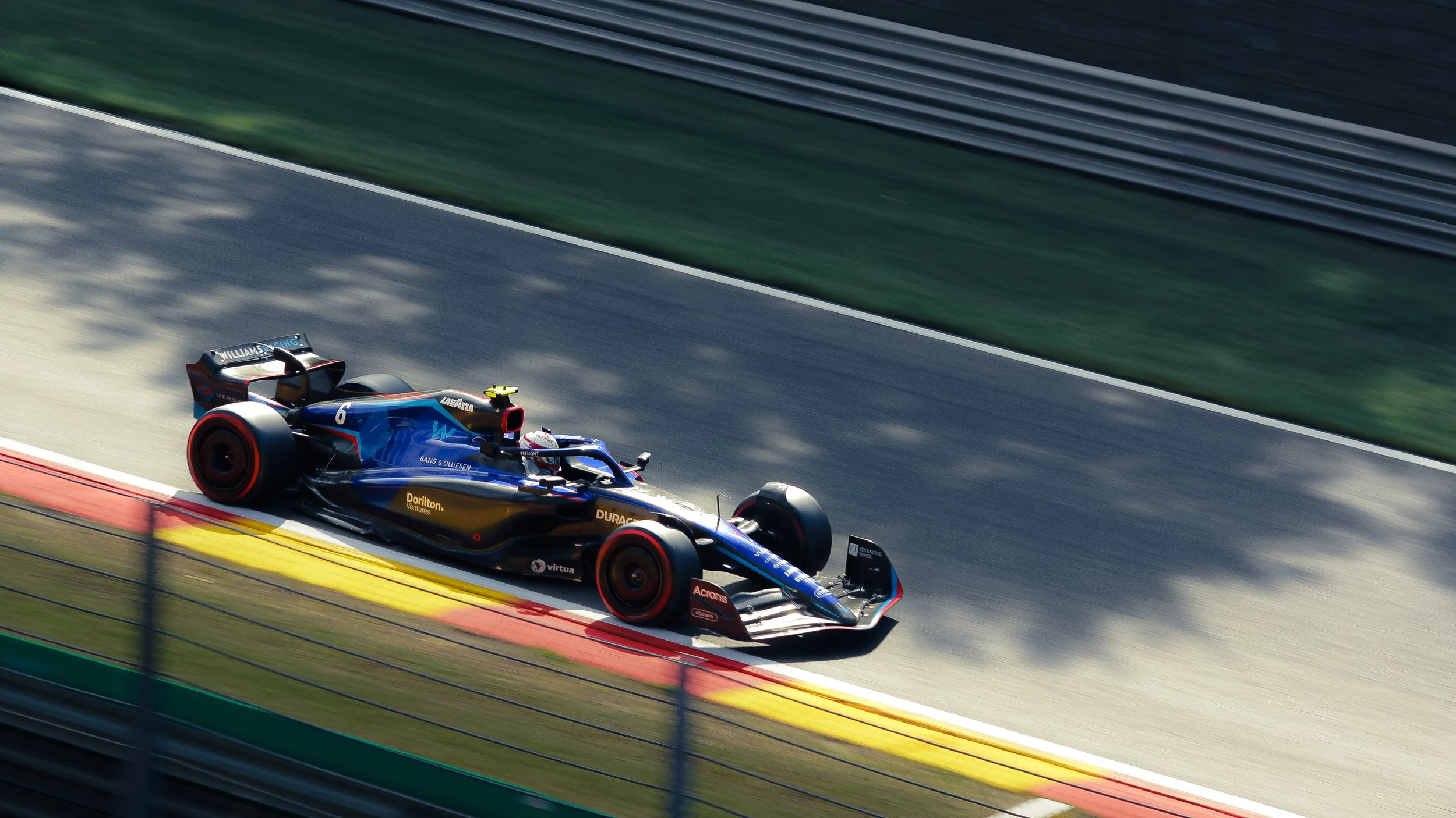 A sleek, blue and black Formula racing car speeding on a race track with red, yellow, and green curbs, with shadows of trees cast across the track.