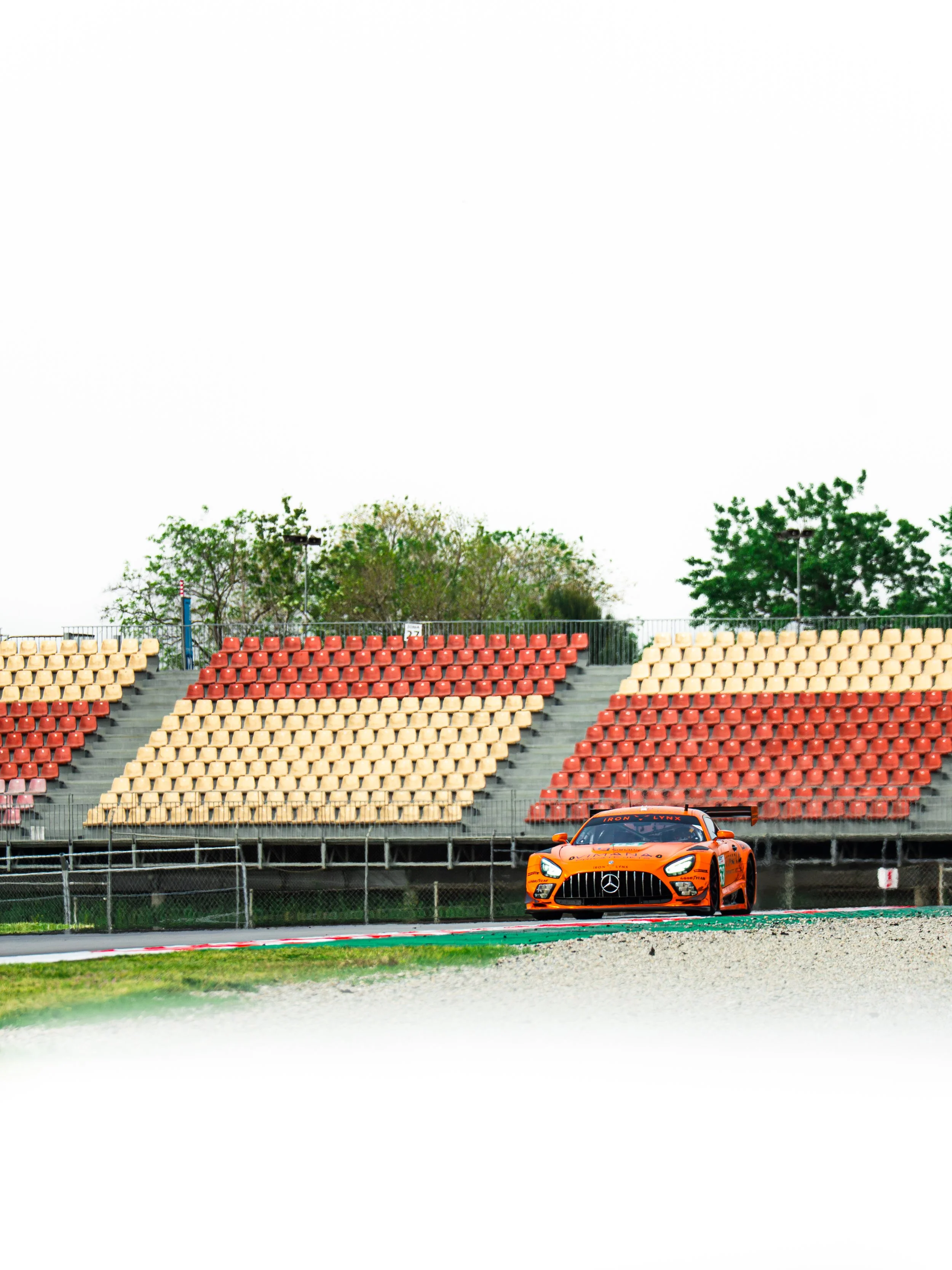 Orange race car on a racing track with empty stadium stands in the background and trees behind the stands.