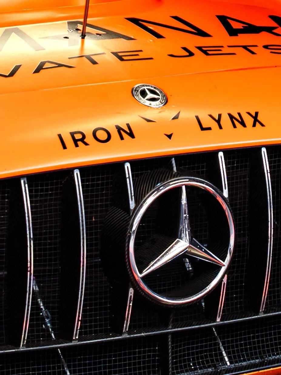 Close-up of the front of an orange Mercedes-Benz race car with the Mercedes logo, grille, and sponsorship decals including 'IRON' and 'LYNX'.