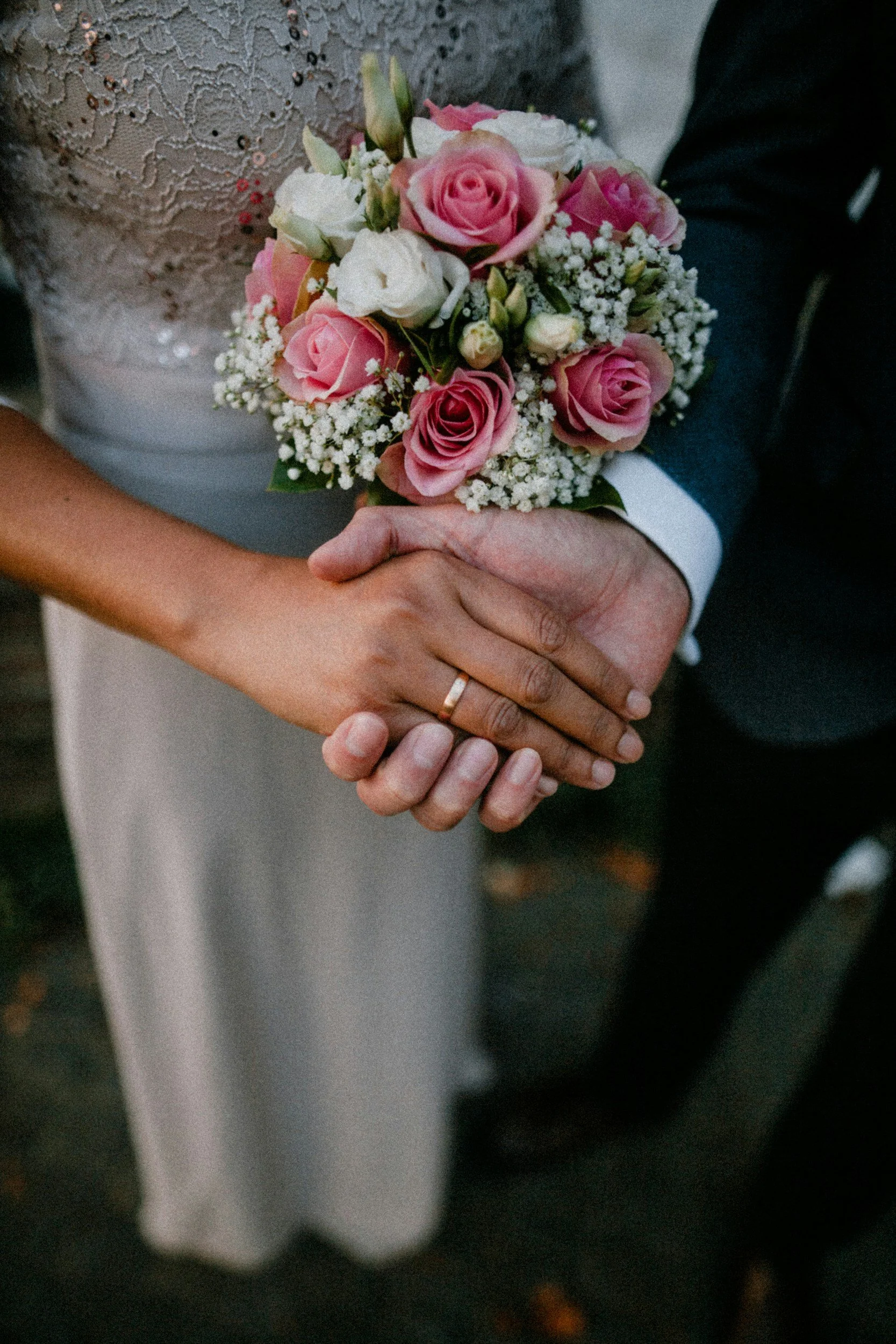 Nahaufnahme eines Brautpaares, das sich die Hände hält. Die Braut trägt einen Ehering, und die Hand hält einen kleinen Blumenstrauß aus rosa und weißen Blumen.