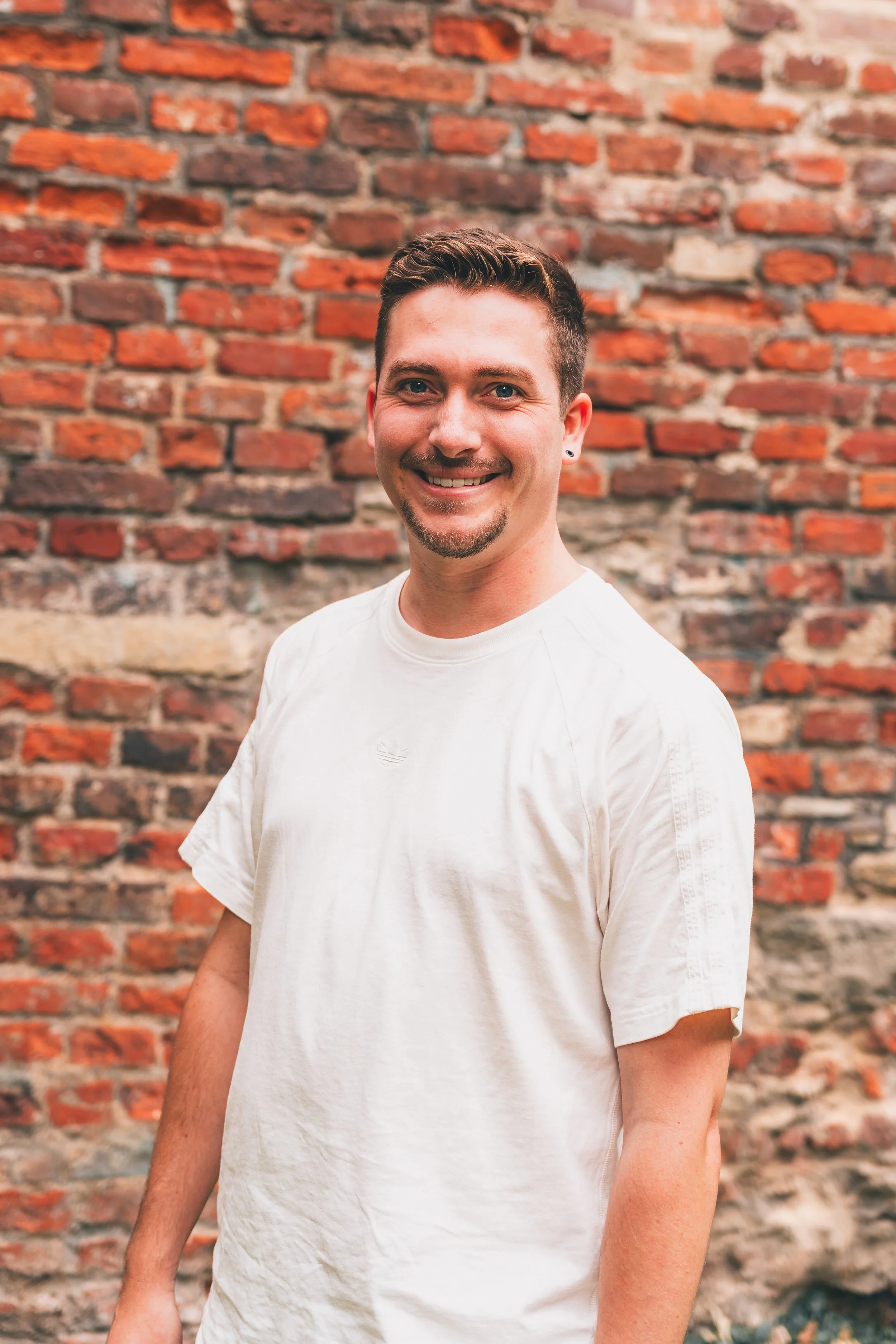 Ein lachender Mann mit dunklem Haar und Kurzhaarschnitt, weißes T-Shirt, vor einer Backsteinmauer.