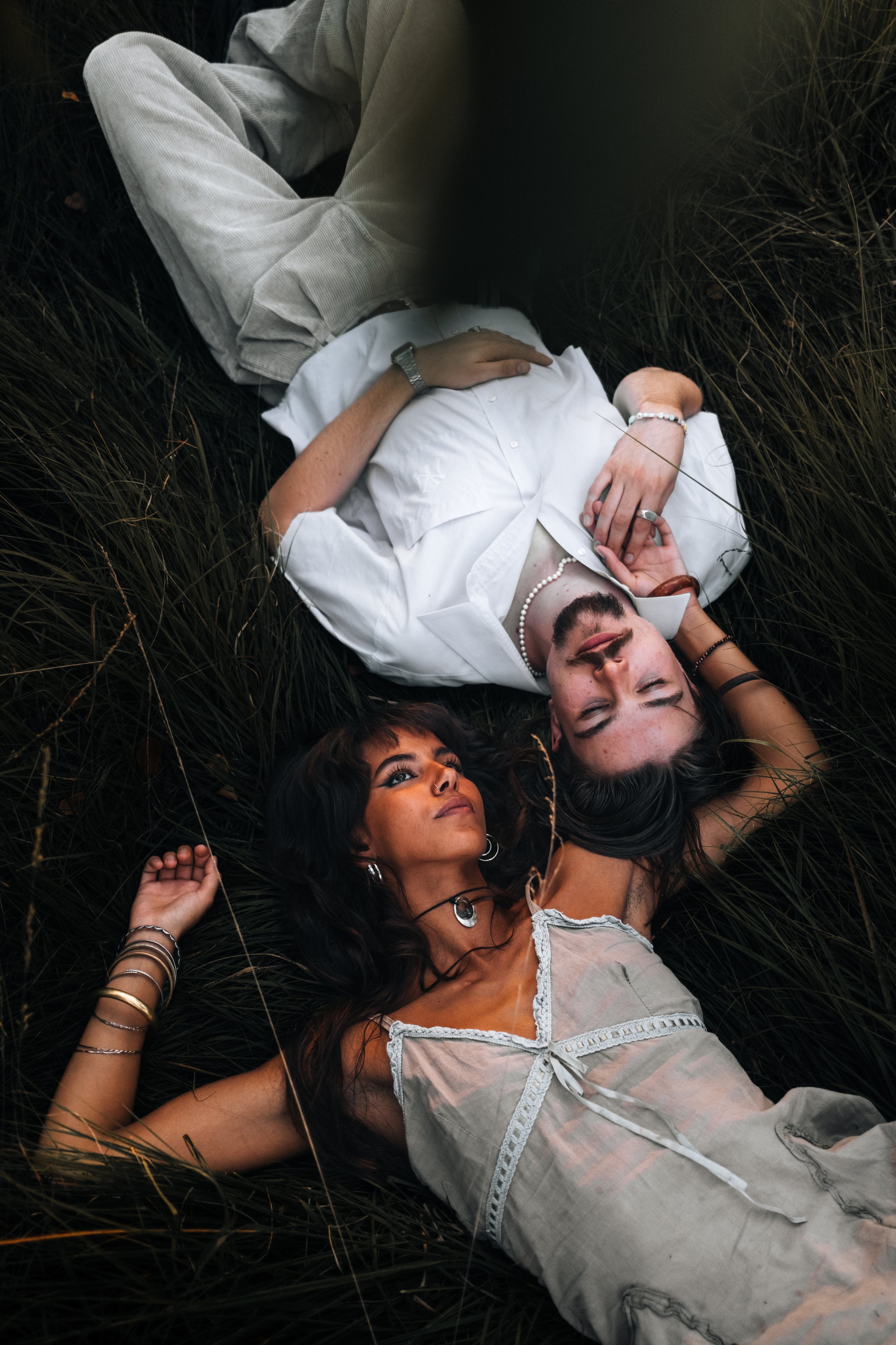 Ein Mann und eine Frau liegen auf hohem Gras, beide mit geschlossen Augen, in einer entspannter Pose, umgeben von natürlicher Umgebung.