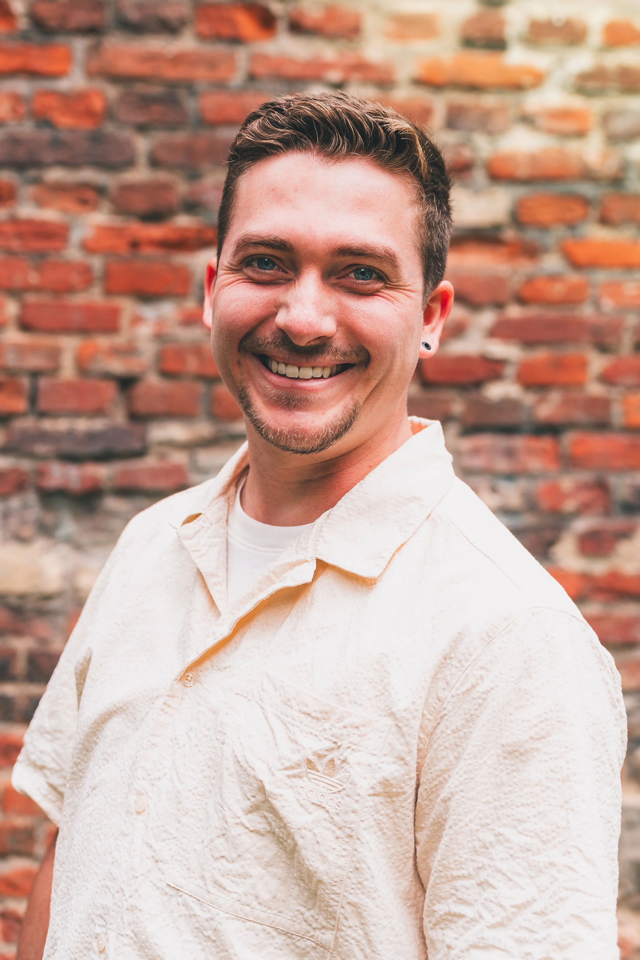 Ein lächelnder Mann mit kurzen braunen Haaren, einem kleinen Barthaar und einem Ohrpiercing, trägt ein cremefarbenes, strukturiertes Hemd vor einer Ziegelmauer.