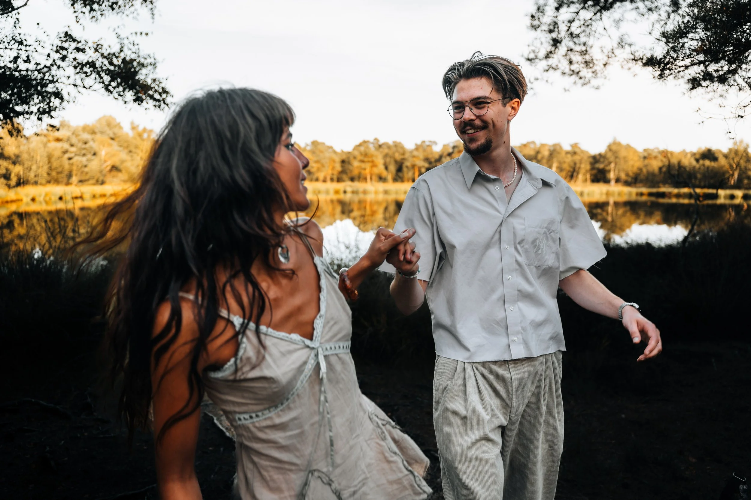 Ein Mann und eine Frau tanzen fröhlich am See bei Sonnenuntergang, umgeben von Bäumen.