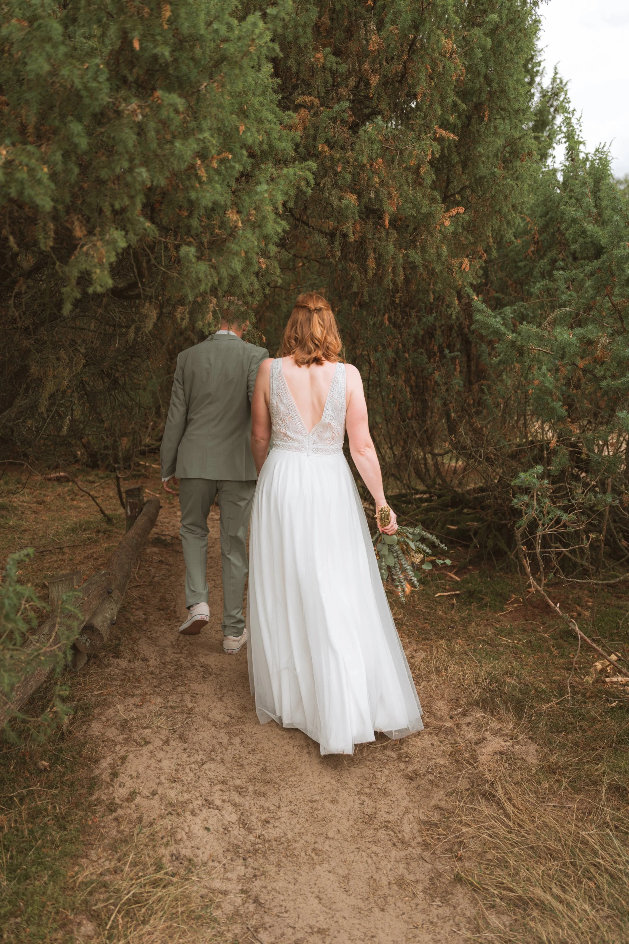 Ein Brautpaar spaziert auf einem Pfad durch einen Wald, die Braut trägt ein weißes Hochzeitskleid, der Bräutigam einen grauen Anzug.