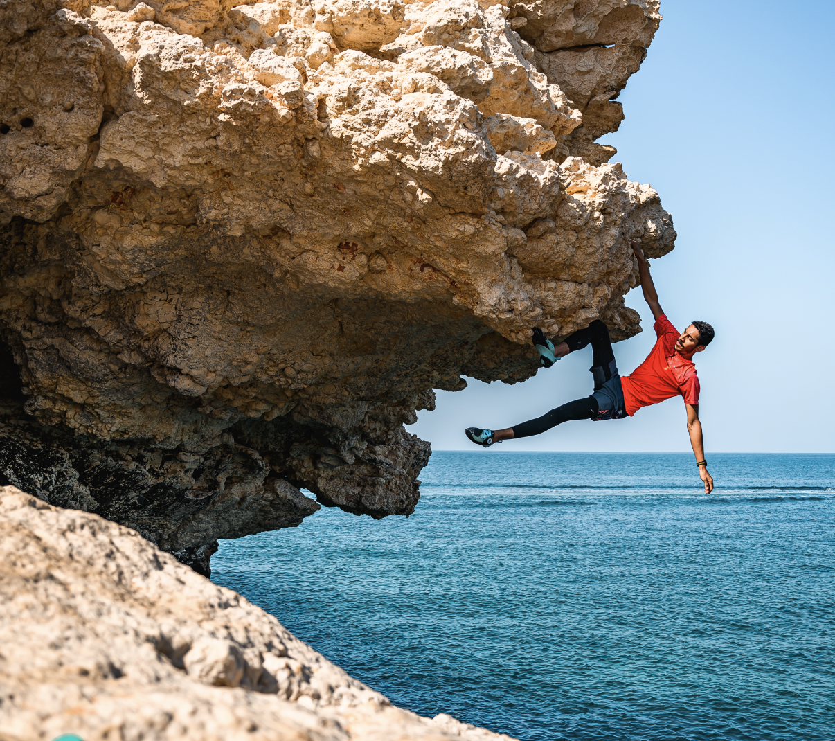 A man in a red shirt and black leggings is hanging from a large rocky overhang over the ocean, holding onto the rock with one hand while extending his body outward.