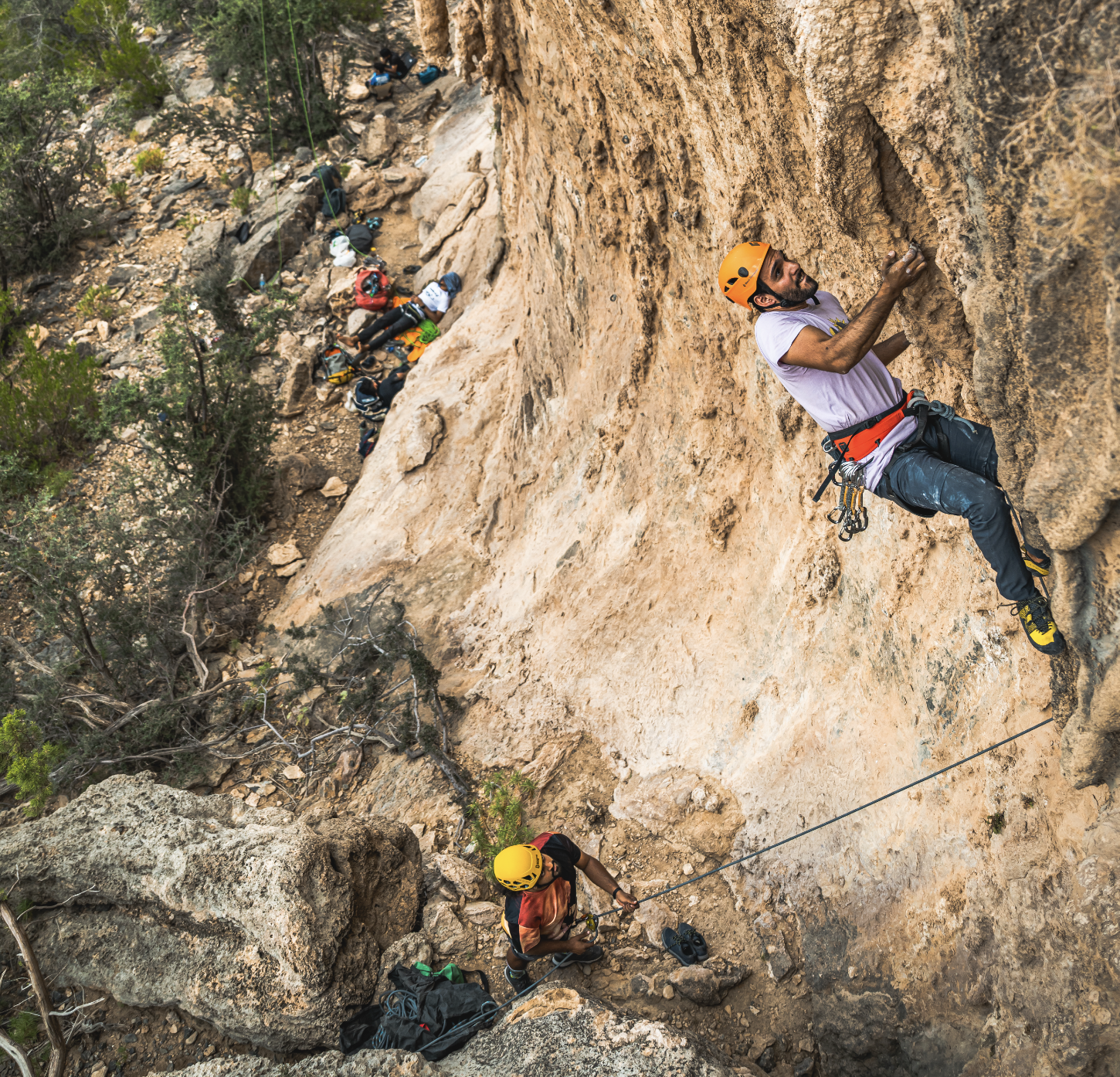 A man rock climbing on a steep cliff, secured with harnesses and ropes, with other climbers and climbing gear below him on the rocky terrain.
