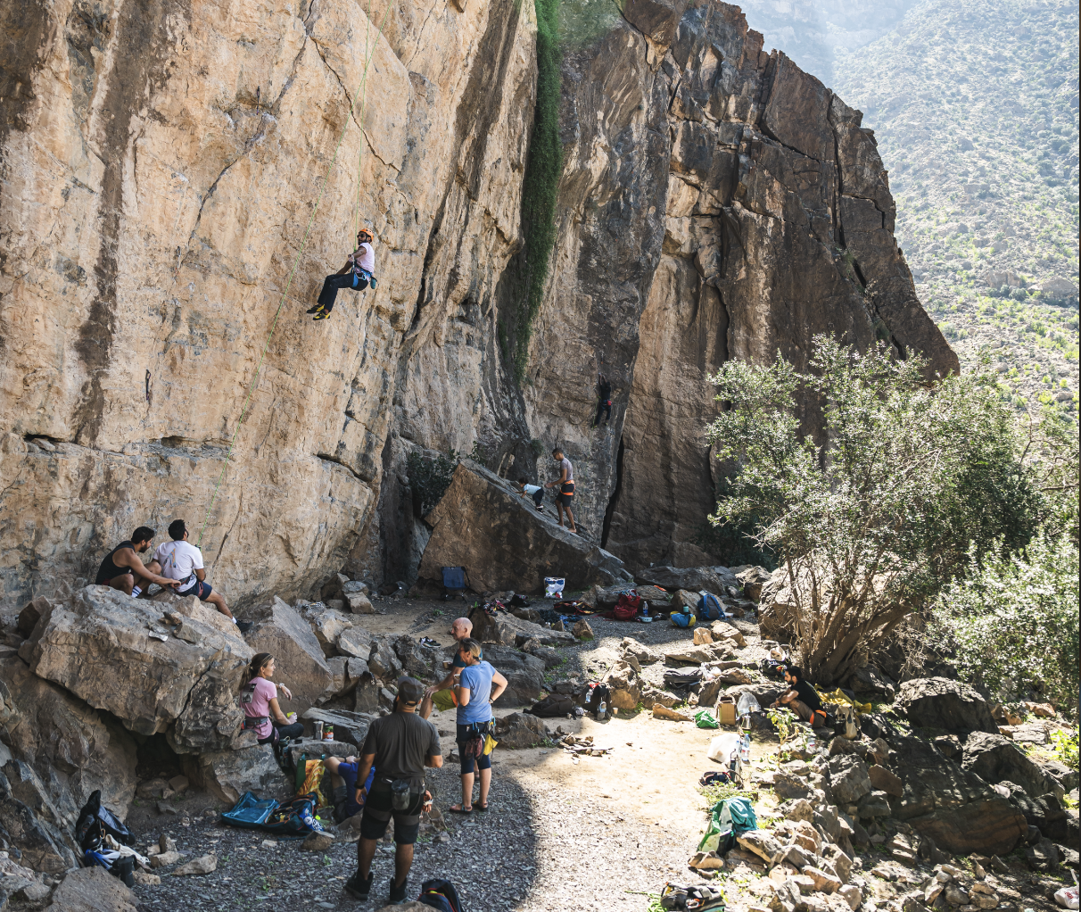 Climbers ascending a steep rock face while others prepare gear and rest at the base in a mountainous outdoor setting.