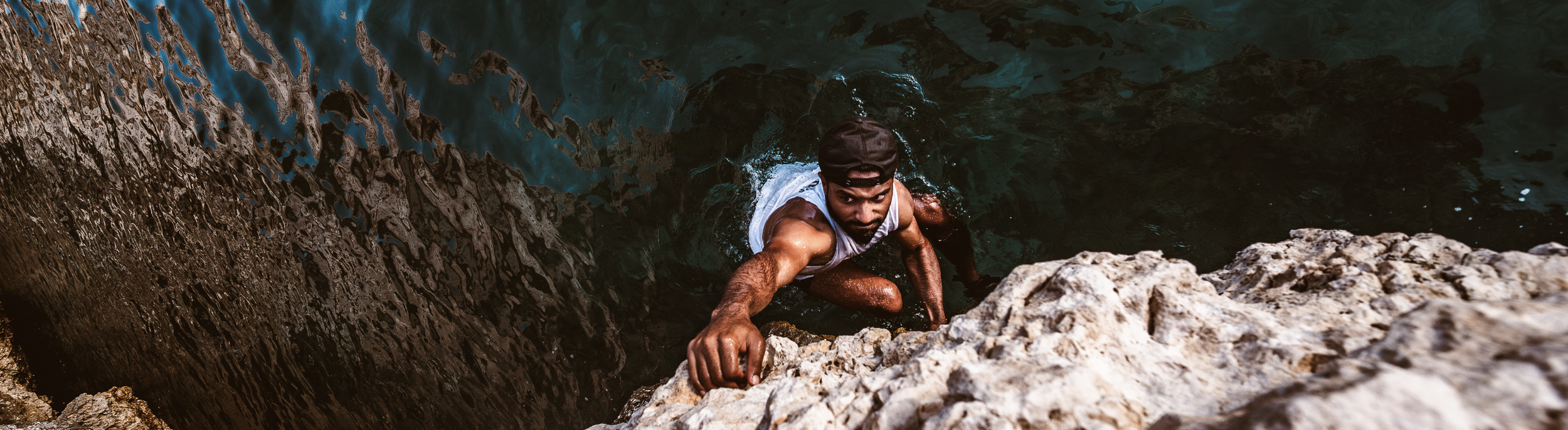 A man climbing out of a natural water hole or pond, looking up at the camera, wearing a black cap and a sleeveless shirt, with wet hair and rocks nearby.
