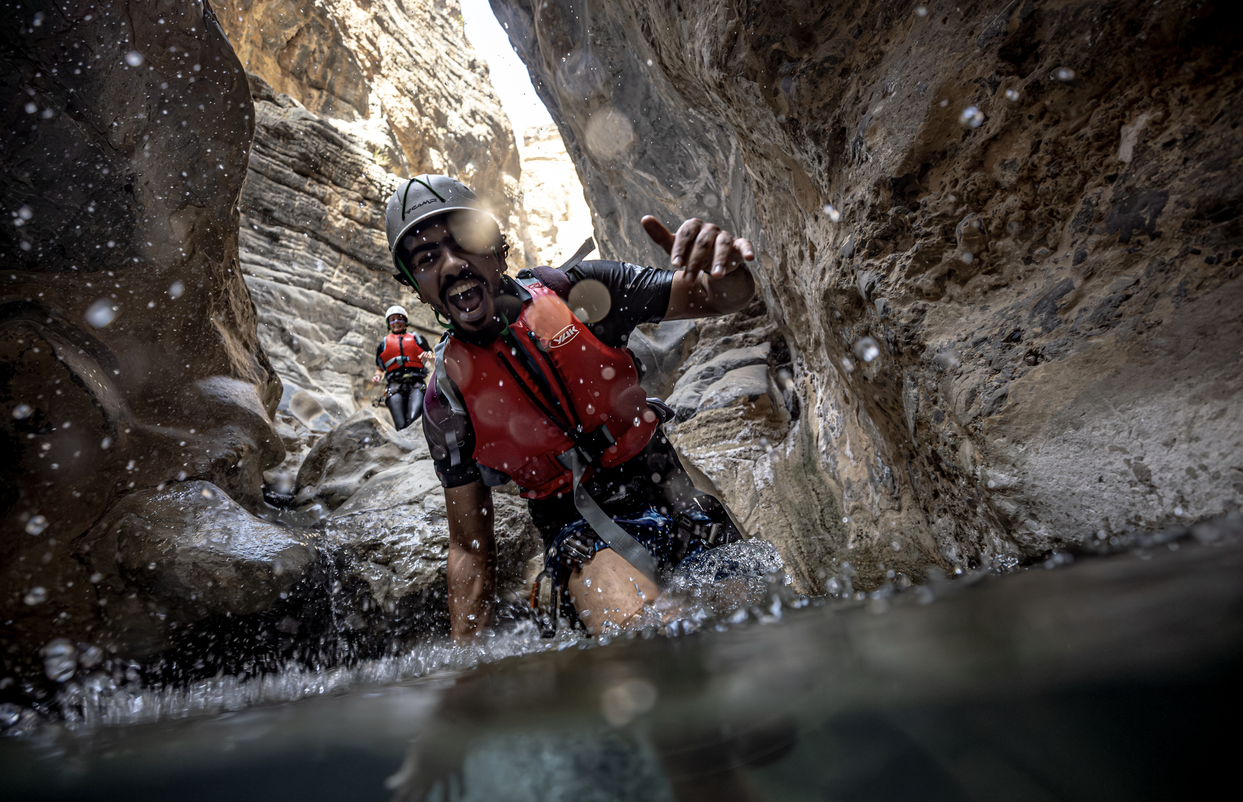 Two people wearing helmets and life jackets are navigating through a narrow canyon with rocky walls, with water splashing around them.