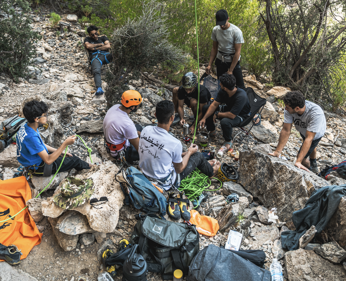 Group of people resting and preparing gear on a rocky, wooded outdoor trail, with climbing and safety equipment, some sitting and some standing.