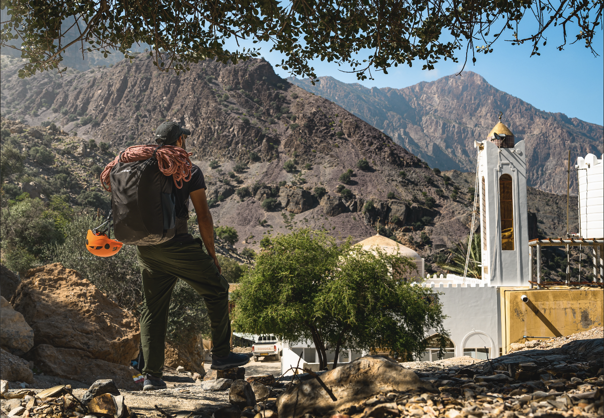 A hiker with a backpack and safety helmet stands on rocky terrain, looking towards a mountain landscape with a white church-style building in the background, under a blue sky with some tree branches at the top.
