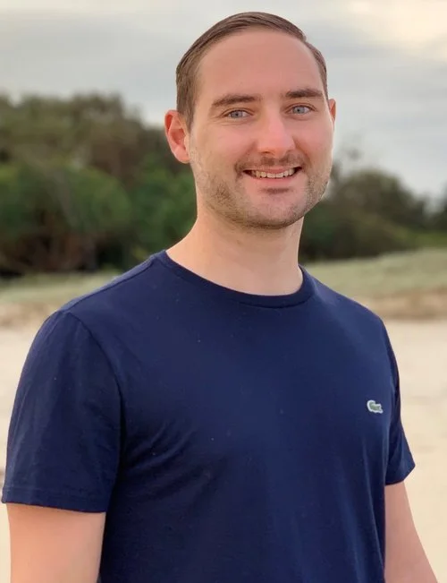 A young man with short hair and a beard, smiling, wearing a navy blue Lacoste t-shirt, standing outdoors on a beach with trees and sand in the background.
