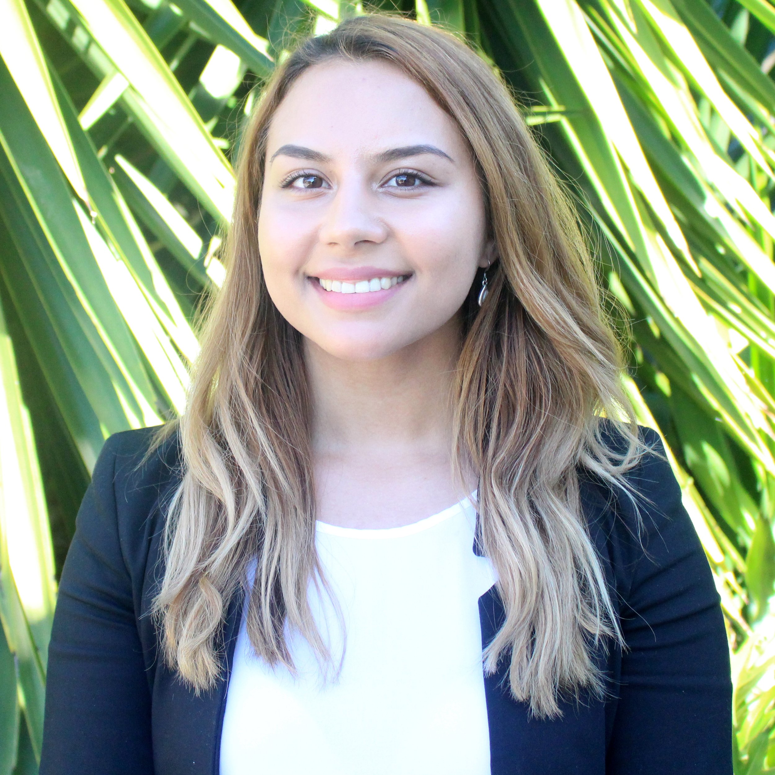 A smiling woman with long wavy blonde hair standing in front of large green foliage.