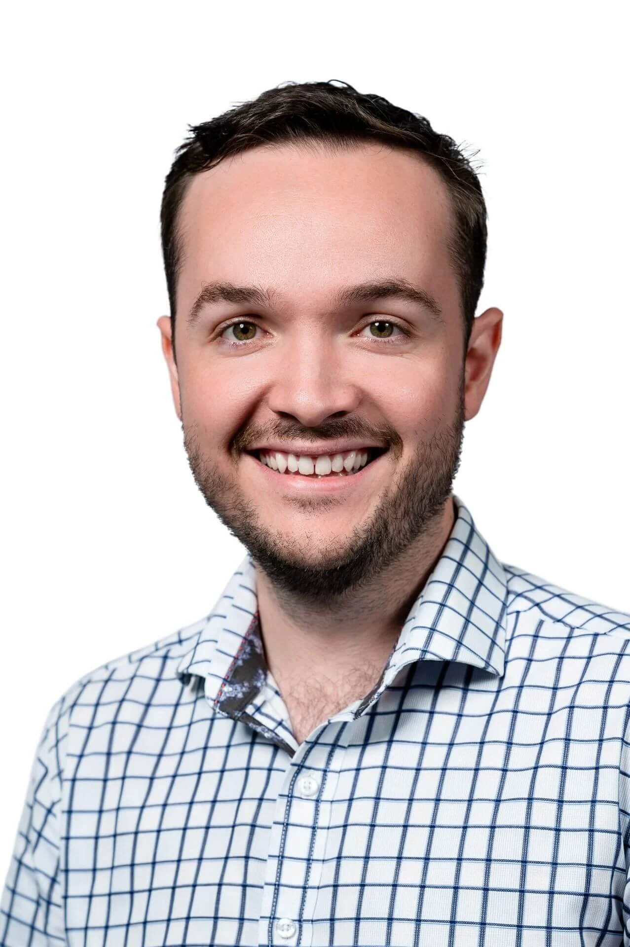 A smiling young man with short dark hair, a beard, and light-colored eyes, wearing a white shirt with blue checks, against a plain white background.