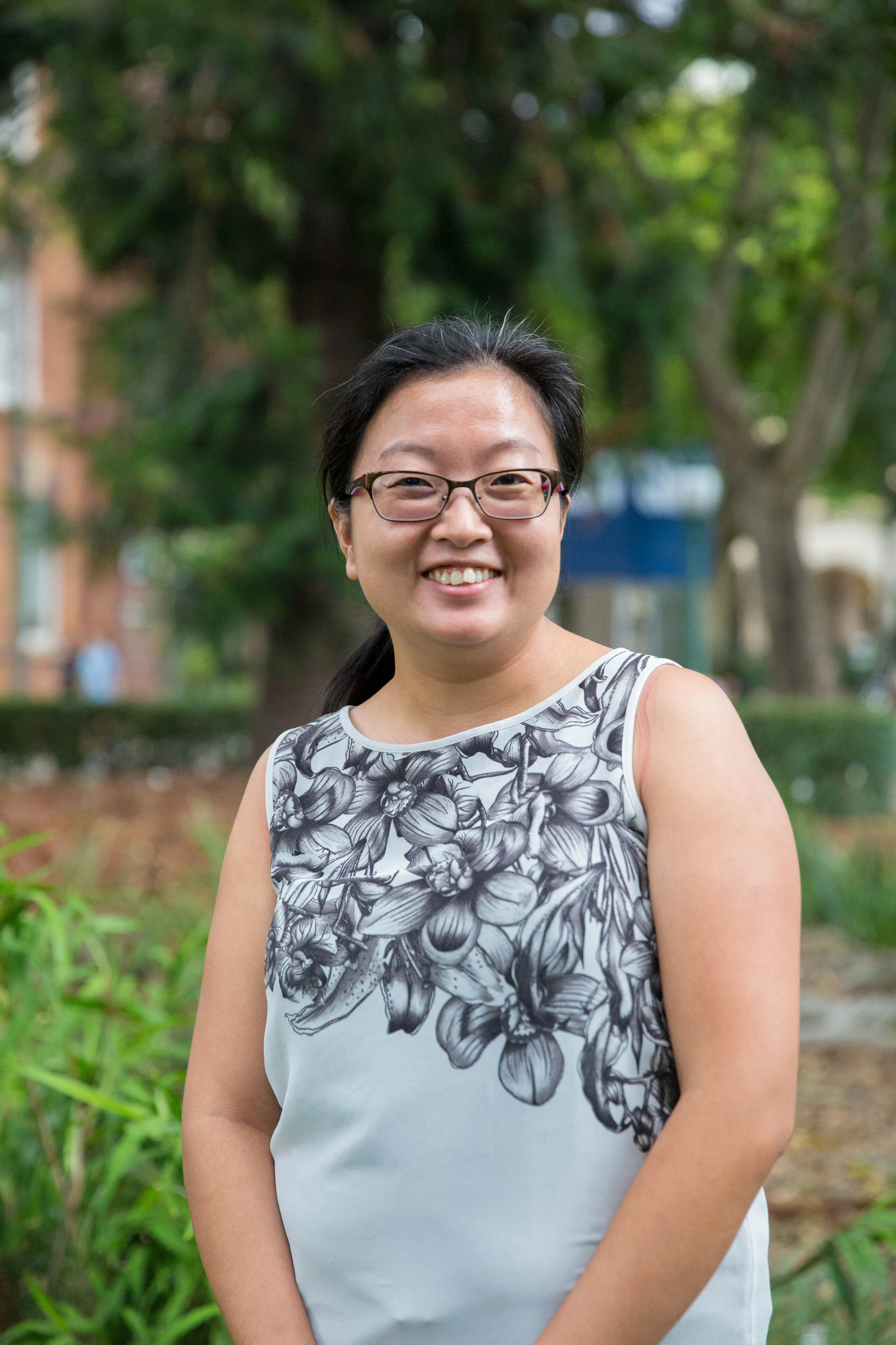 A smiling woman with glasses and dark hair tied back, wearing a sleeveless top with a floral black and white pattern, standing outdoors in a green park with trees and bushes in the background.