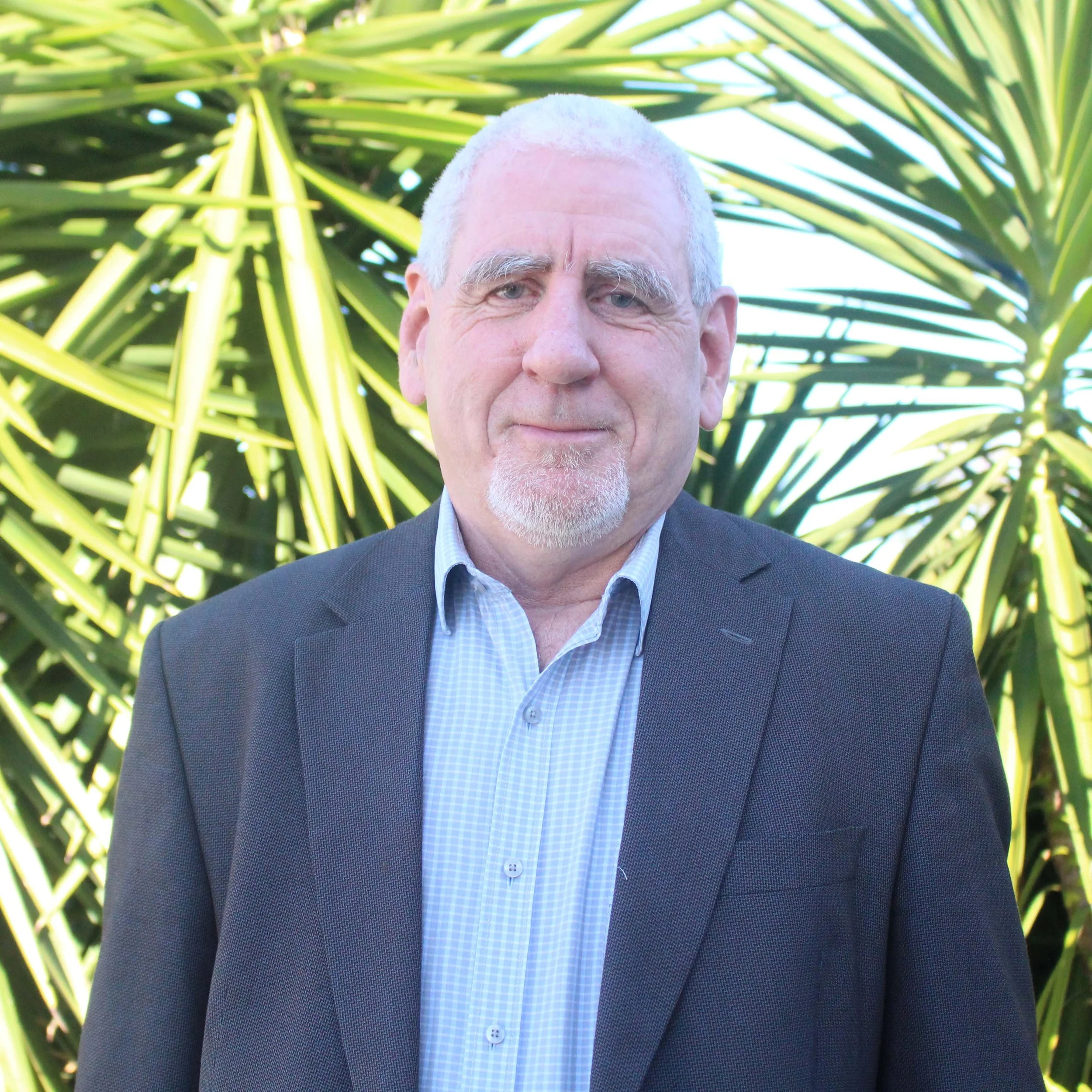 Close-up portrait of a smiling mature man with white hair and beard, wearing a dark blazer and light blue checked shirt, standing outdoors in front of green tropical plants.